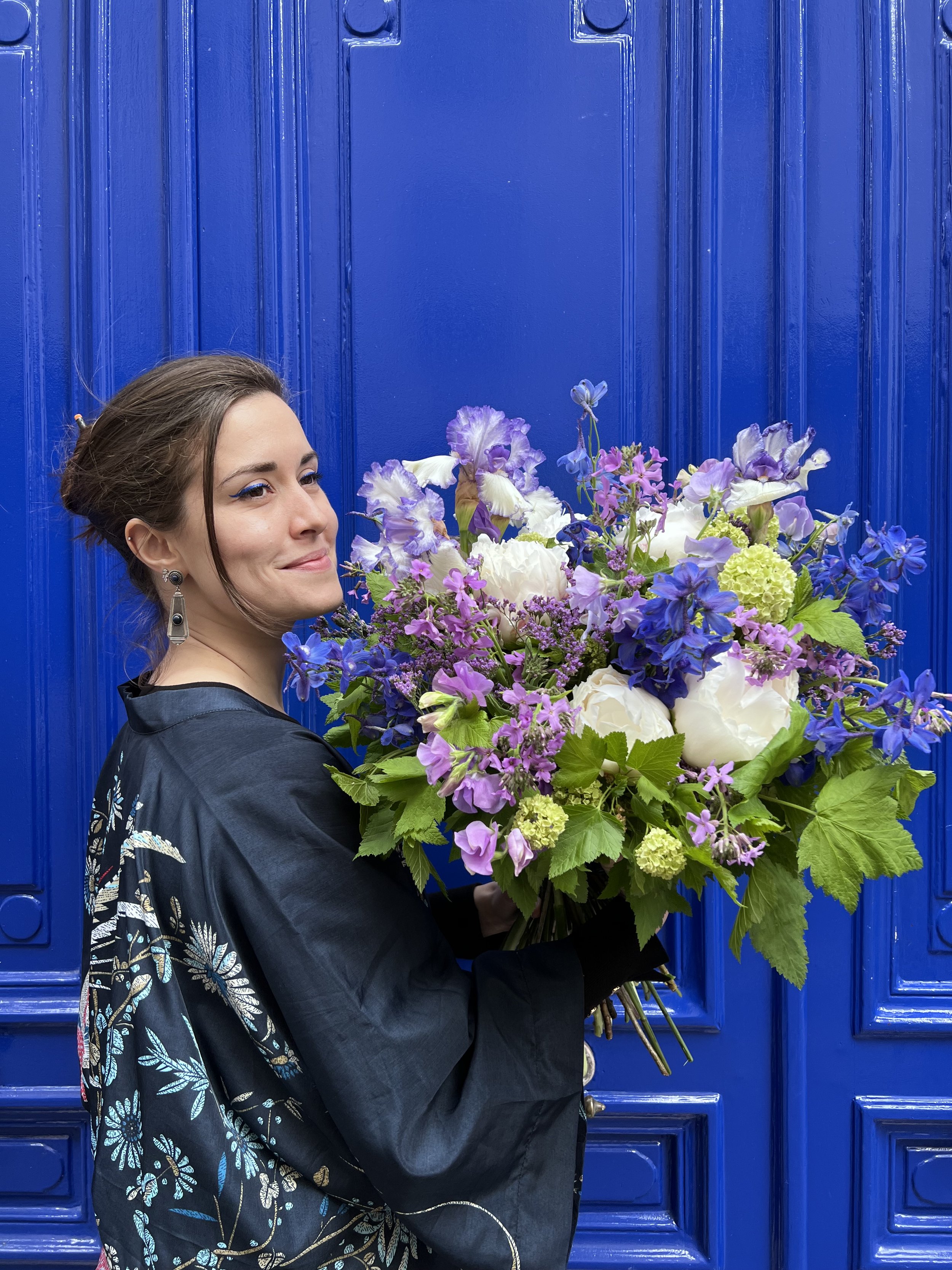 Une femme tenant un grand bouquet de fleurs colorées, avec un fond de porte bleue.