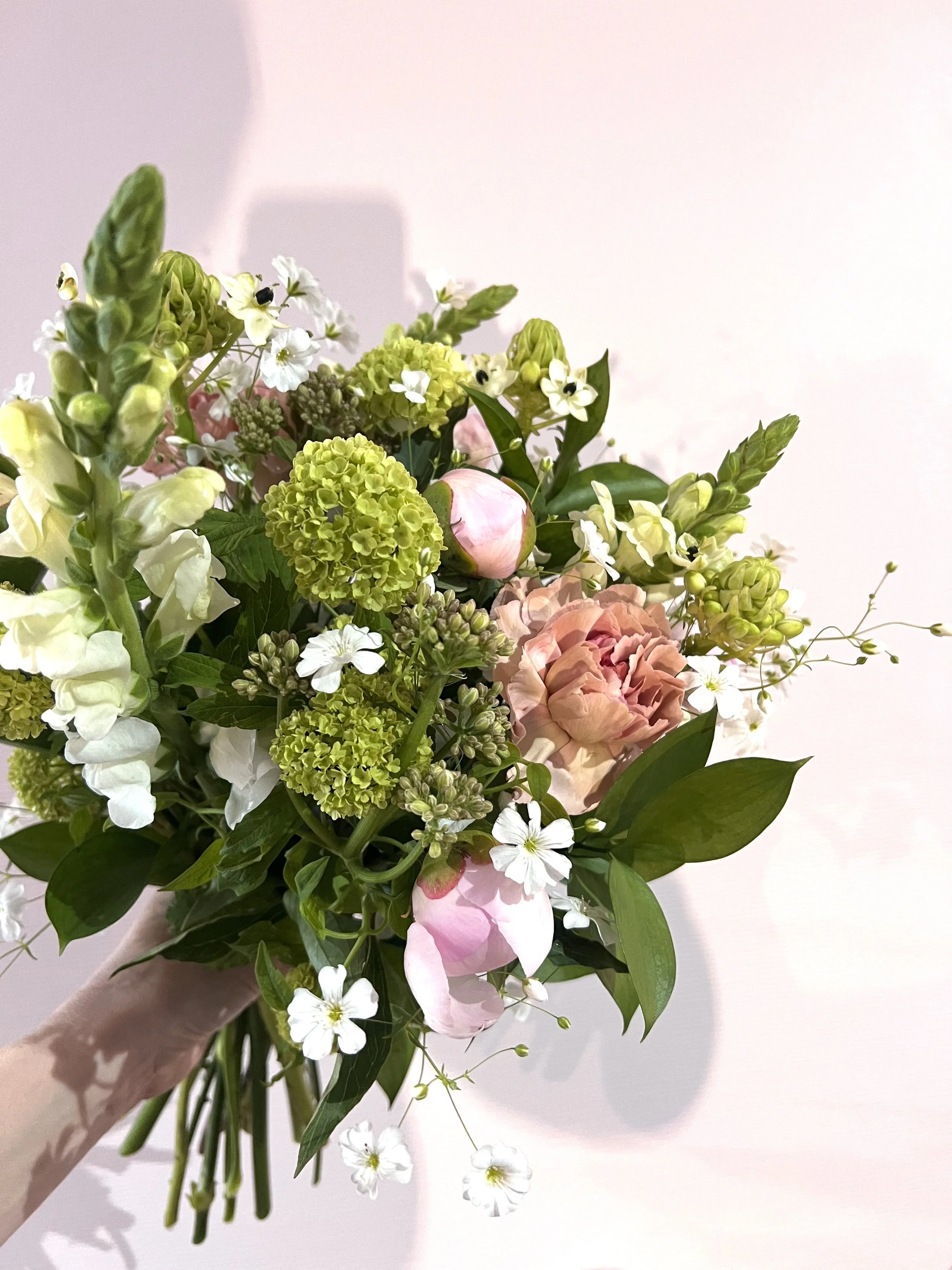 Un bouquet de fleurs colorées avec des roses, des hortensias, des petites fleurs blanches et des feuilles vertes, tenu par une main
