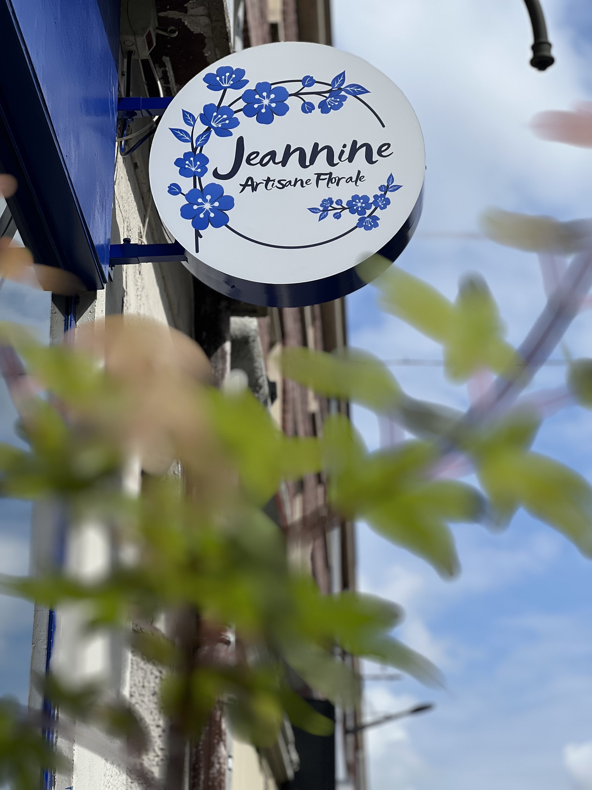 Pancarte ronde blanche avec des fleurs bleues et le texte 'Jeannine Artisane Florale' écrit en noir, fixée à un mur, avec un ciel bleu en arrière-plan.