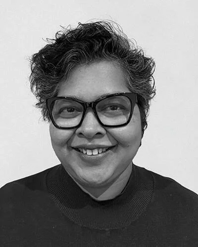 A person with short, curly hair wearing glasses and a dark sweater, smiling against a plain background.