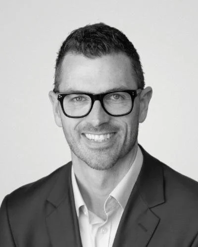 Black and white headshot of a man wearing glasses, a suit, and a light-colored shirt, smiling at the camera.