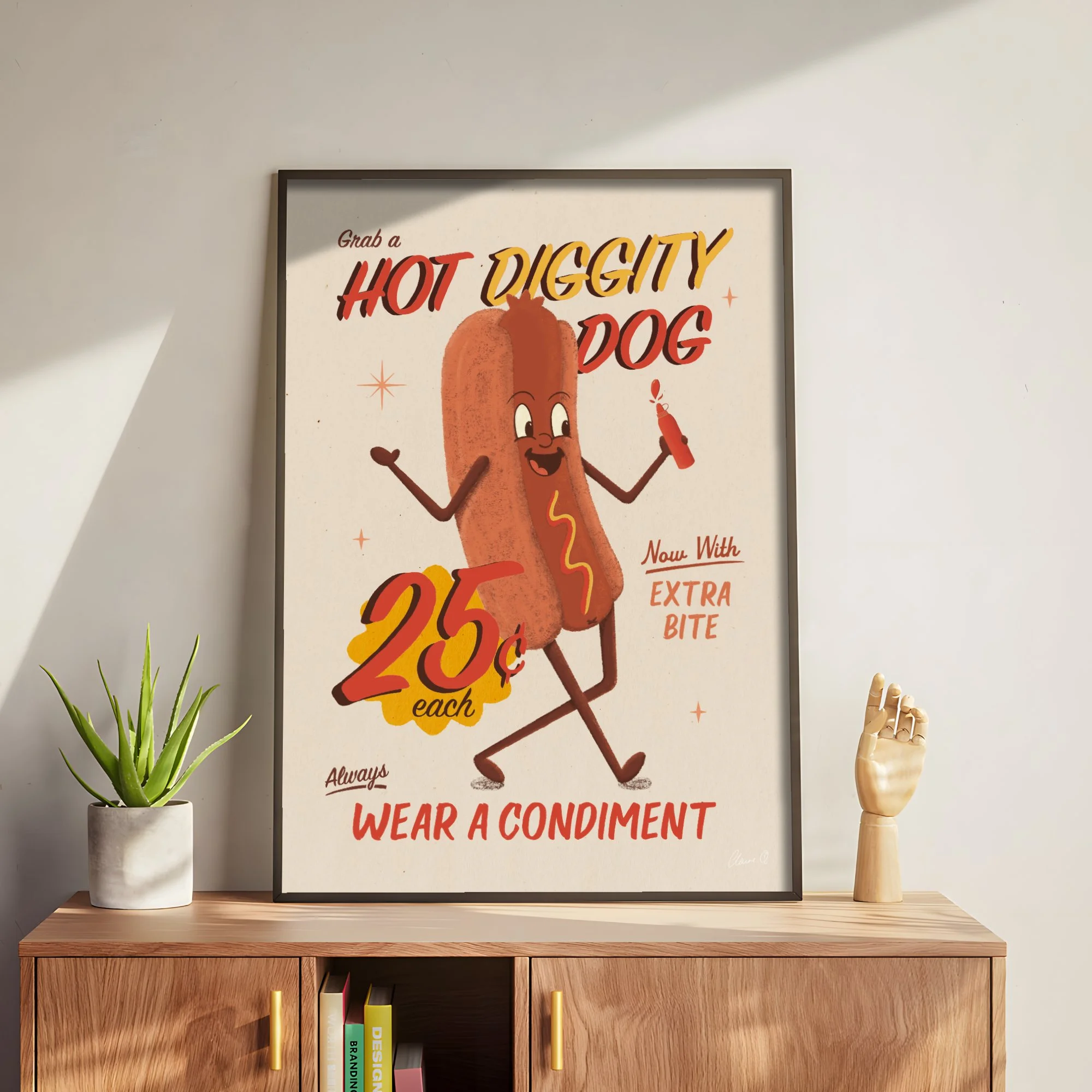 Decorative framed poster on a wall with a retro cartoon hot dog character, promoting wearing a condiment as part of the headline, alongside a plant in a white pot on a wooden table, and a wooden hand mannequin.
