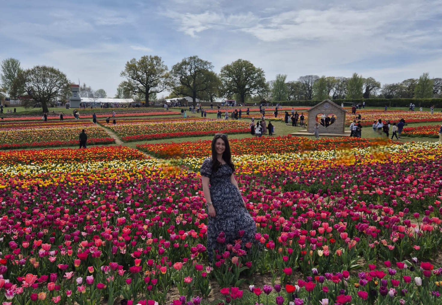 Founder and graphic designer, Claire, in a long floral dress smiling in a colorful tulip field with many people, trees, and a small wooden heart-shaped structure in the background.
