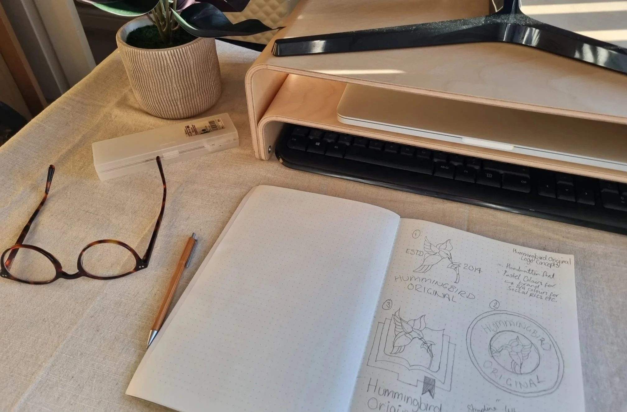 A desk with a potted plant, a pair of glasses, a notepad with sketches and notes, a pen, and a computer keyboard visible in the image.