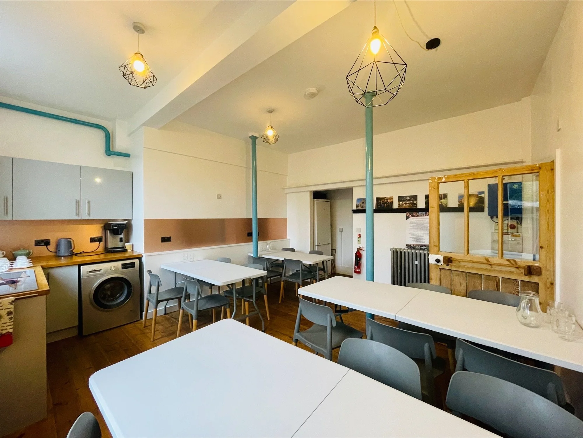 A beautiful professional workshop space with high ceilings, large white tables, lots of seats, and a kitchenette with washing machine, sink and kettle.