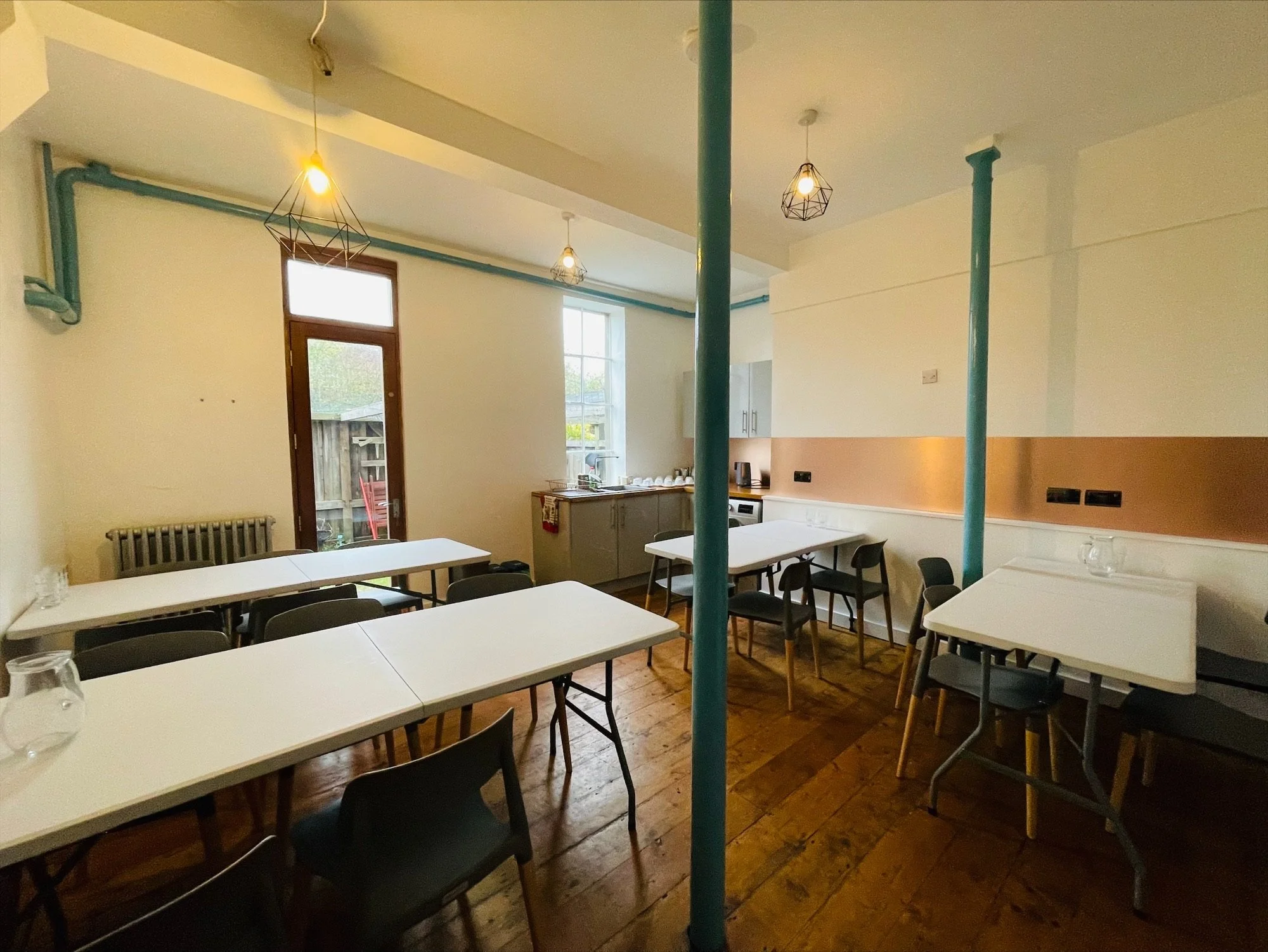 An interior meeting room with several white tables, black chairs, a kitchen area, wood flooring, pendant lights, and a large window.
