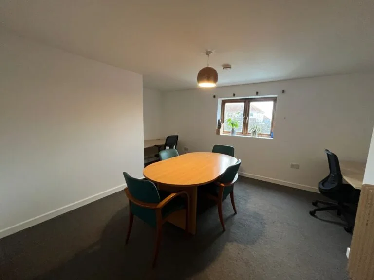 Our affordable office space for hire, white walls, with a large brown conference table, nice window overlooking the garden, and desks in the corners of the room with green chairs next to them.