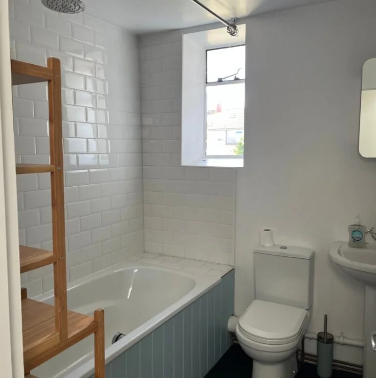 A clean white bathroom with a bath and shower, a window, a toilet and sink and a wooden shelf.