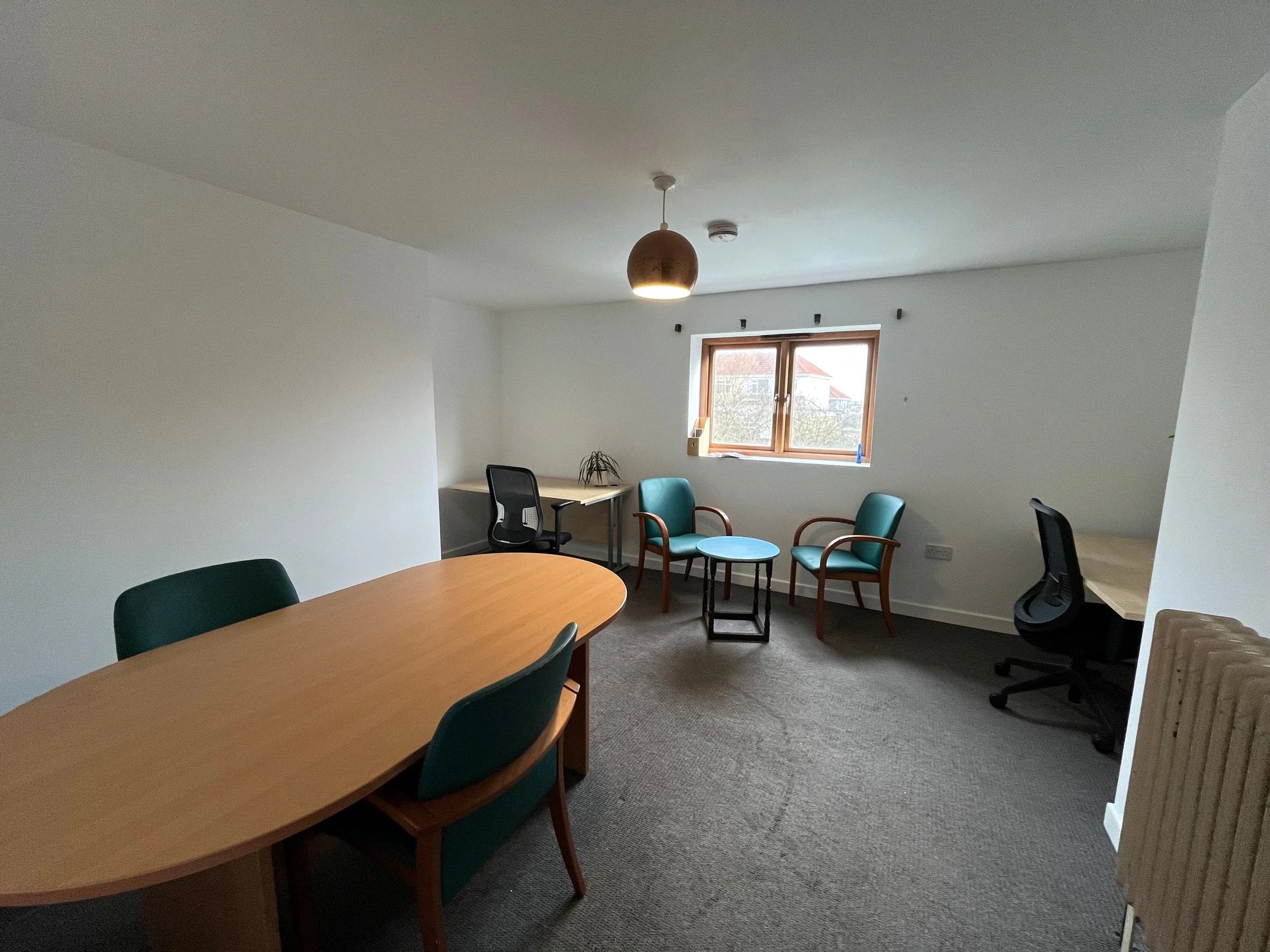 Affordable office room for hire with a wooden meeting table, green chairs, two small armchairs, two desks, and a nice big window.
