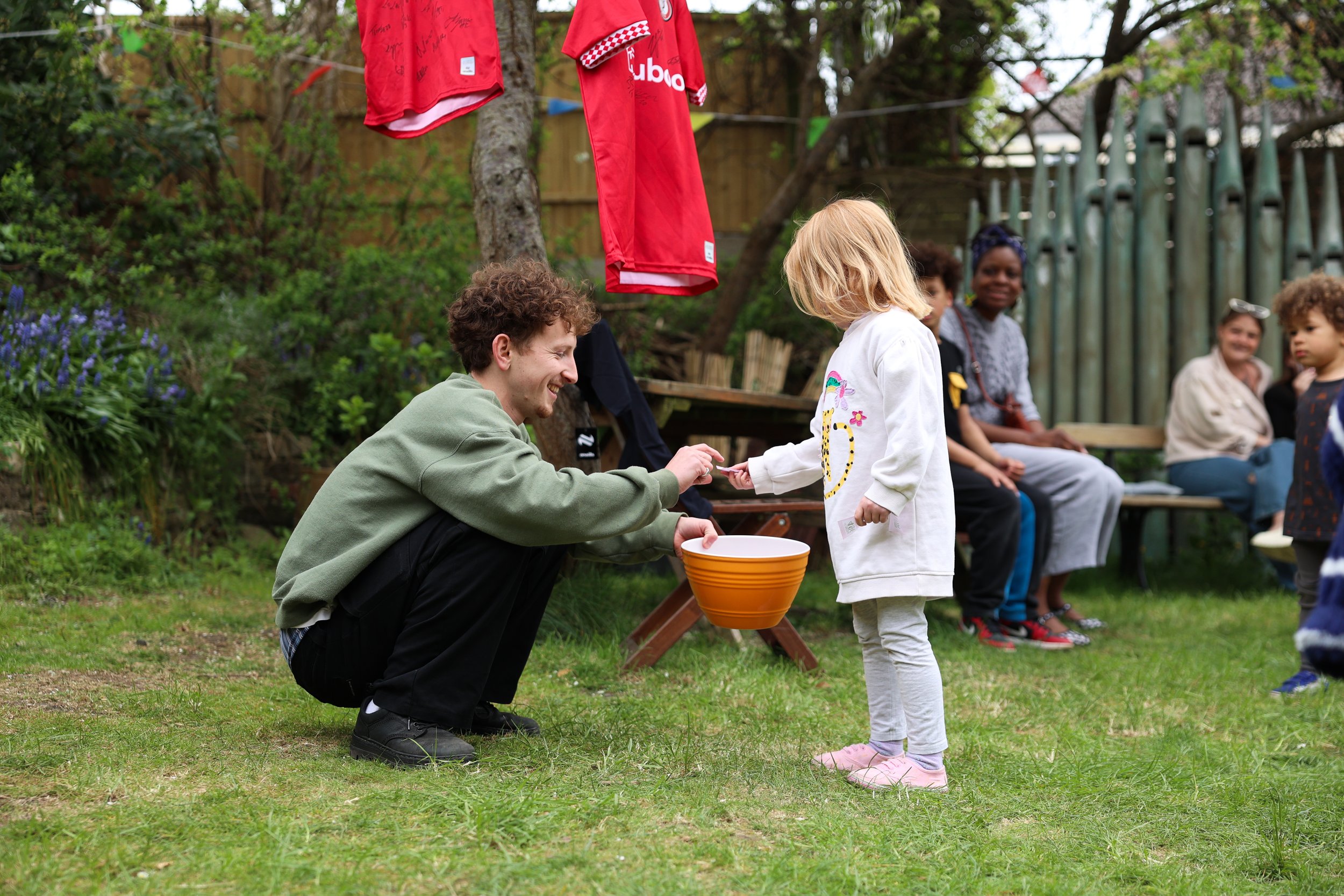 A man kneeling and smiling as he hands a piece of paper to a young girl in a white sweater in our lovely sun filled garden space.