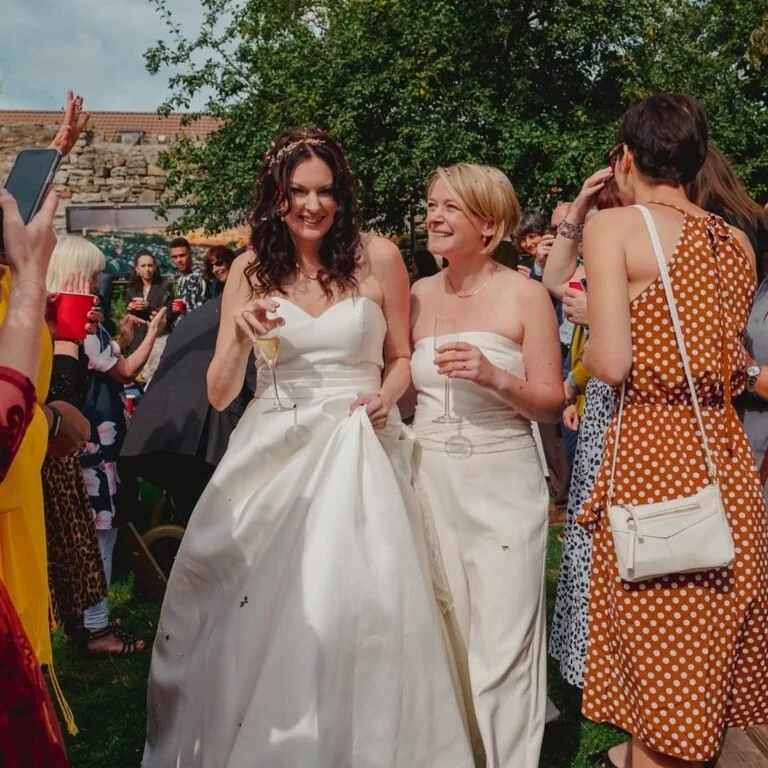 A beautiful gay wedding, two women in white wedding dresses smiling and holding glasses of champagne in our garden surrounded by friends and family.