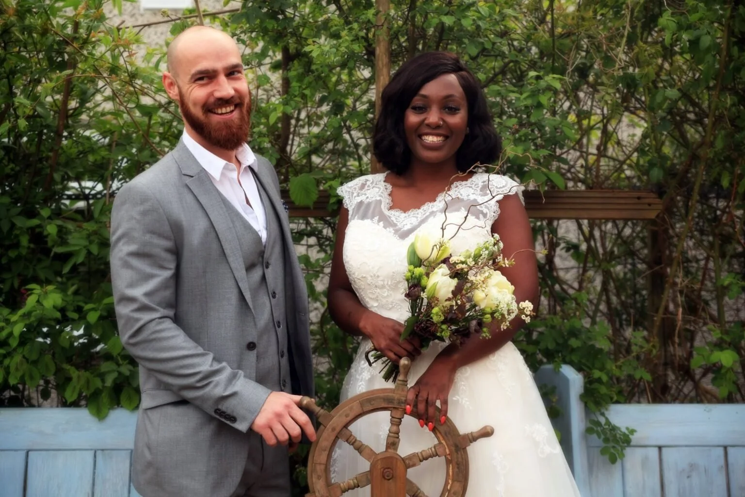 A couple taking wedding photos in our beautiful garden. The groom is white and bald in a grey suit, the bride is black with long black hair and holding a beautiful bouquet of flowers. They're holding a ships wheel between them.