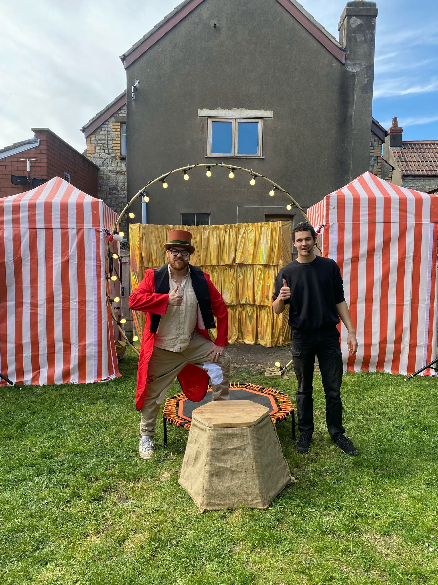 Two men in front of a  circus-themed backdrop with striped tents, fairy lights, and a gold curtain. One man is dressed in a red ring leader coat and the other all in black.