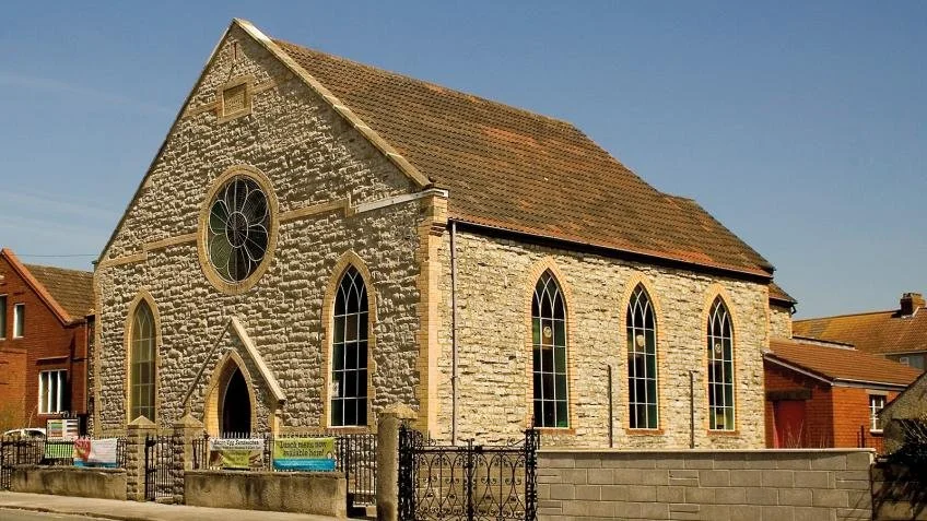An old Methodist building, a small stone church with Gothic-style arched windows, a circular stained glass window with a flower pattern, and a brown roof, on a lovely sunny day.