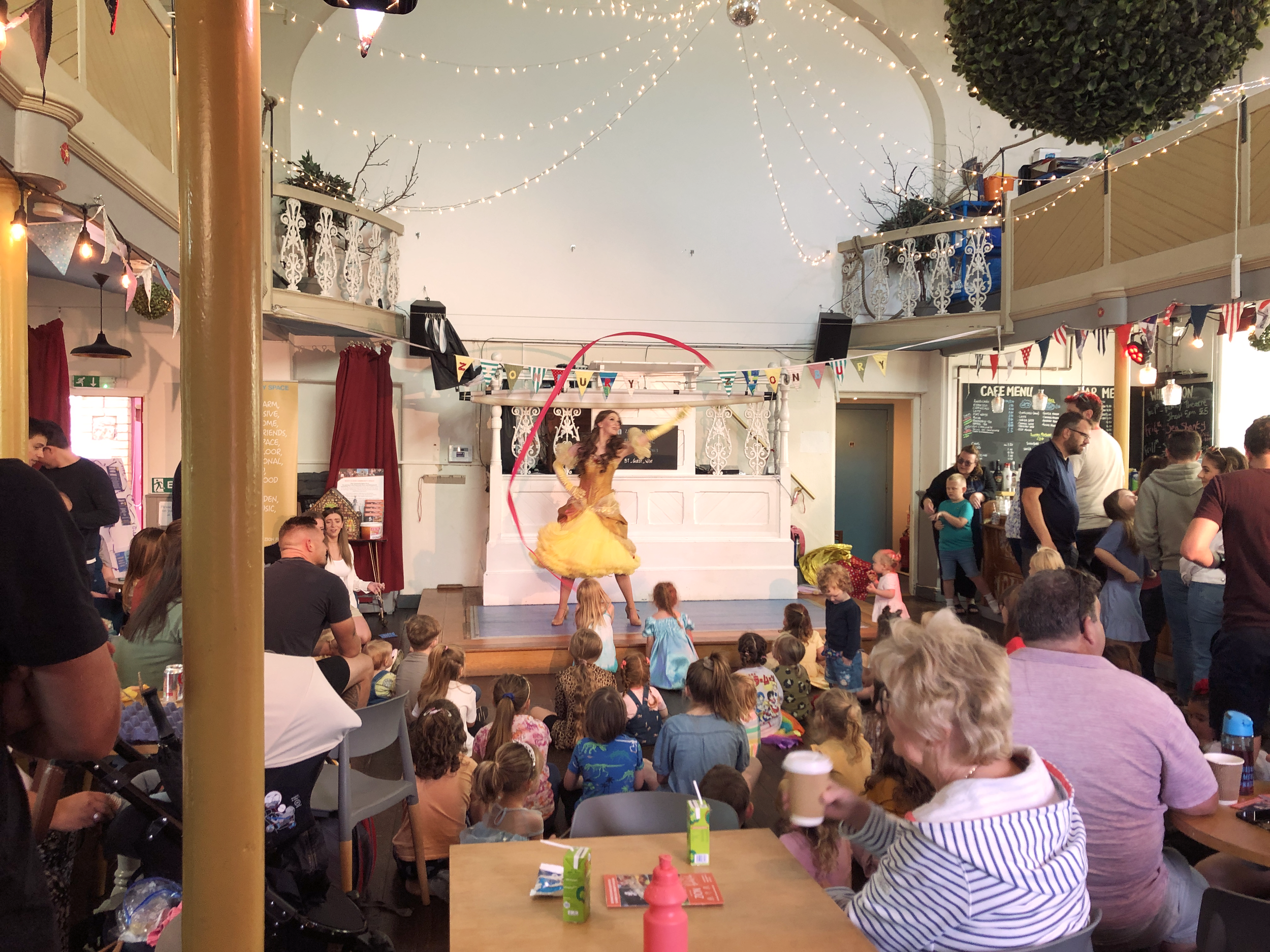A kids performer in a yellow dress spins ribbons on stage, surrounded by children and their adults watching the performance. Decorated with string lights and banners, the café is gorgeous and full of local people having a great time!