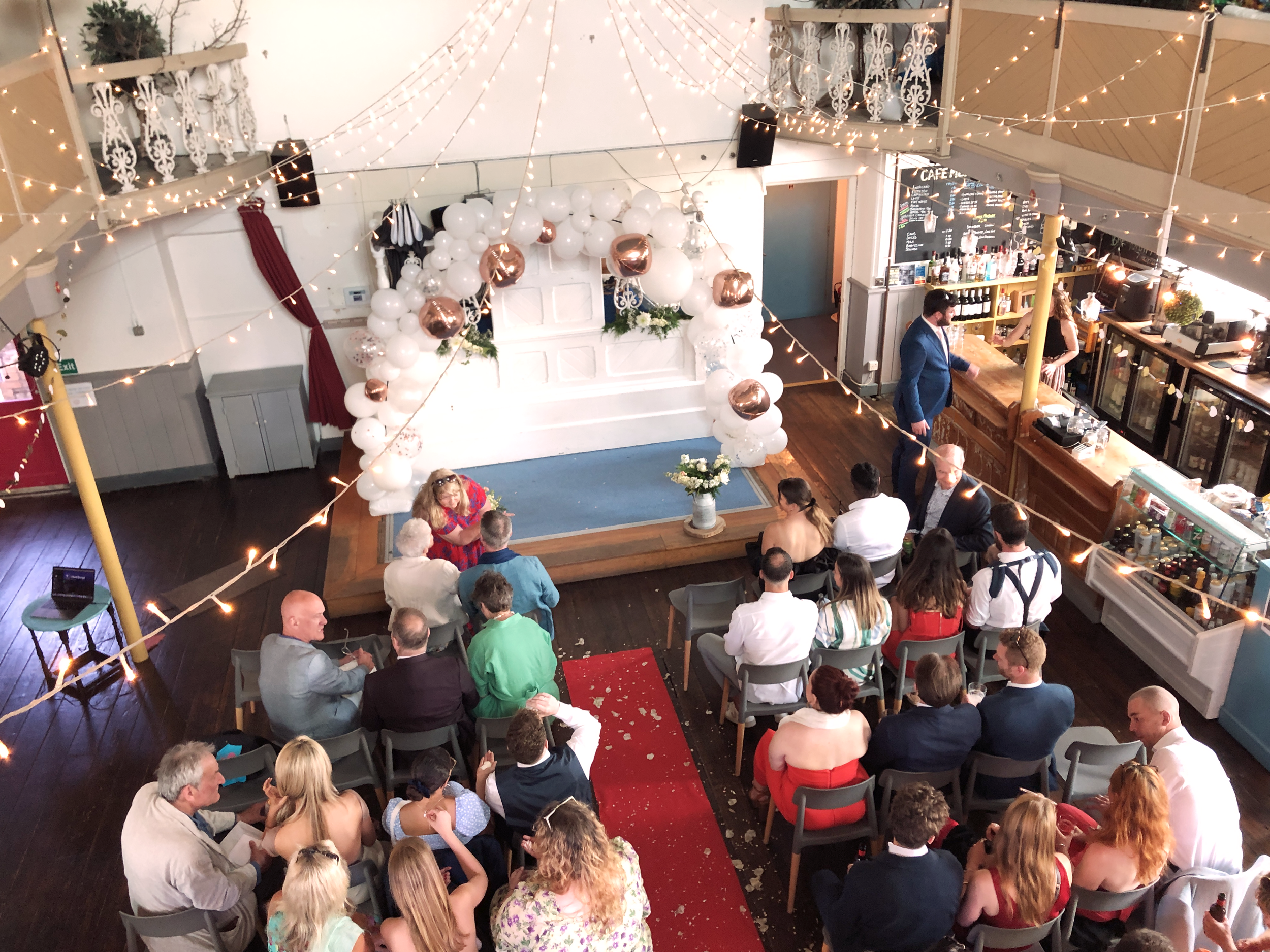 Our café is transformed into a beautiful wedding venue with balloons and string lights. People are sat in their Sunday best waiting for the couple to appear.