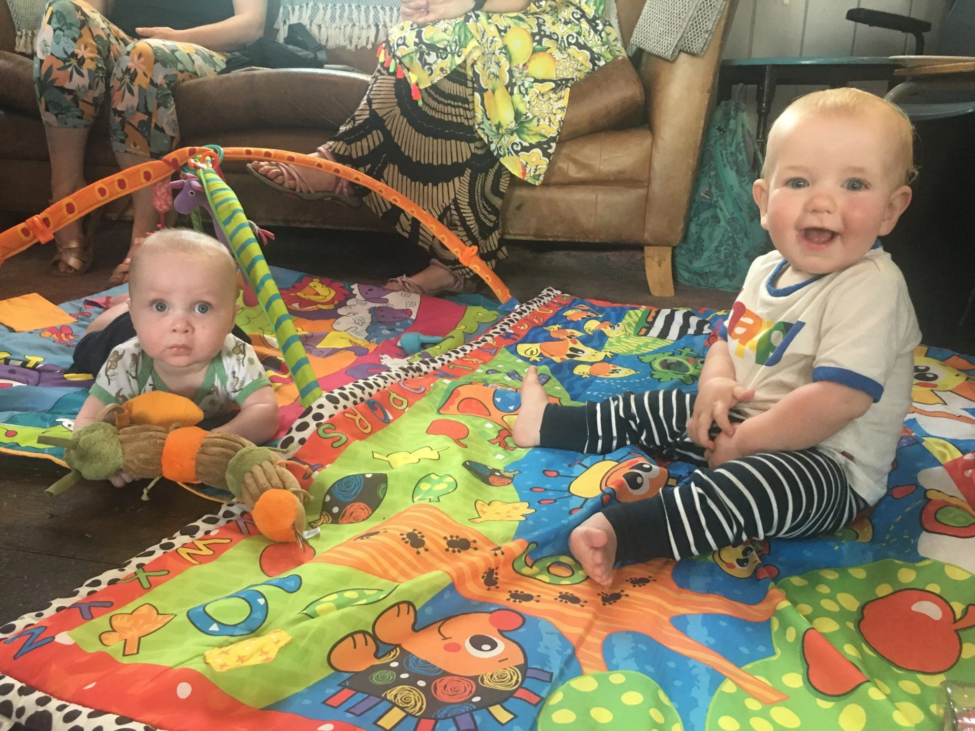 Two white smiling babies play on a colourful, kids blanket with plush toys and large sensory play things. One of the babies is smiling happily at the camera, the other looks shy.