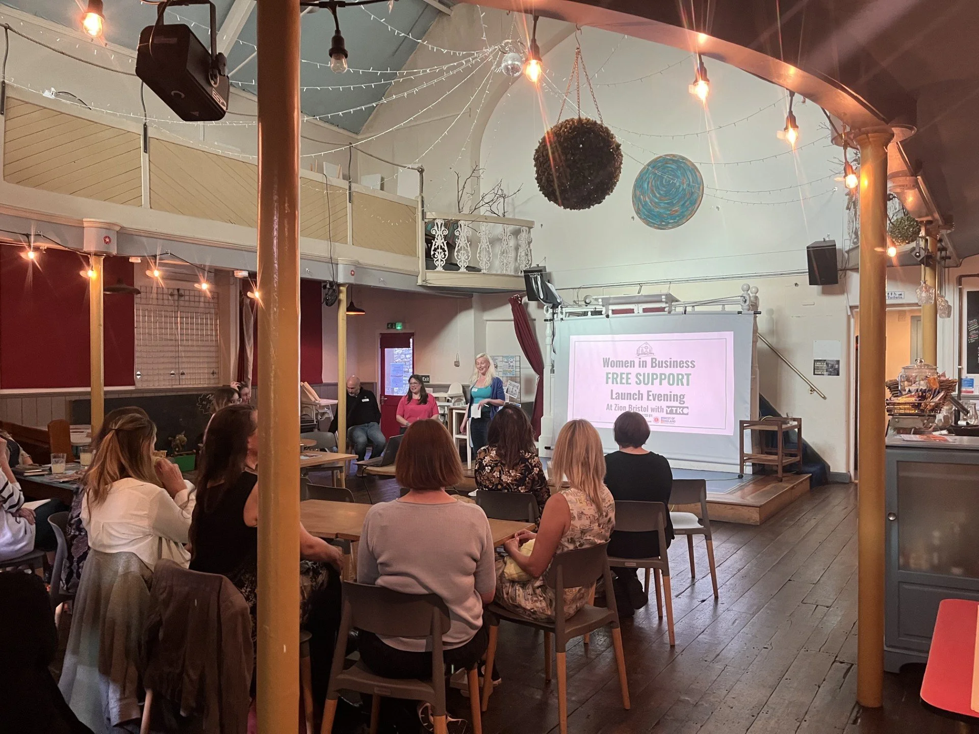 Women attending an evening of 'Women in Business Free Support' at Zion Bristol. The cafe looks beautiful with its high, decorated ceiling, and string lights. A speaker stands near the screen, and attendees are seated at tables listening attentively.
