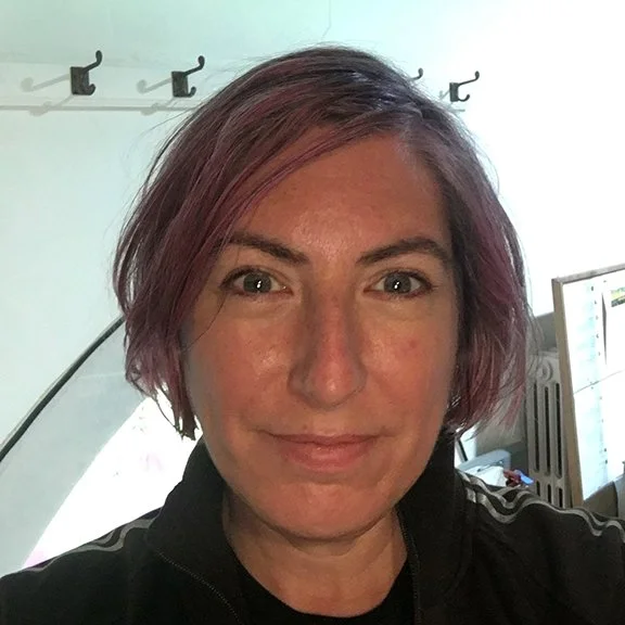 A woman with short purple hair taking a selfie indoors, she has a lovely smile and looks confident.