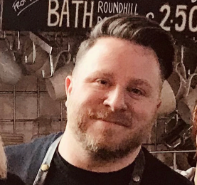 A man with short hair and a beard smiling, wearing a black shirt and an apron.