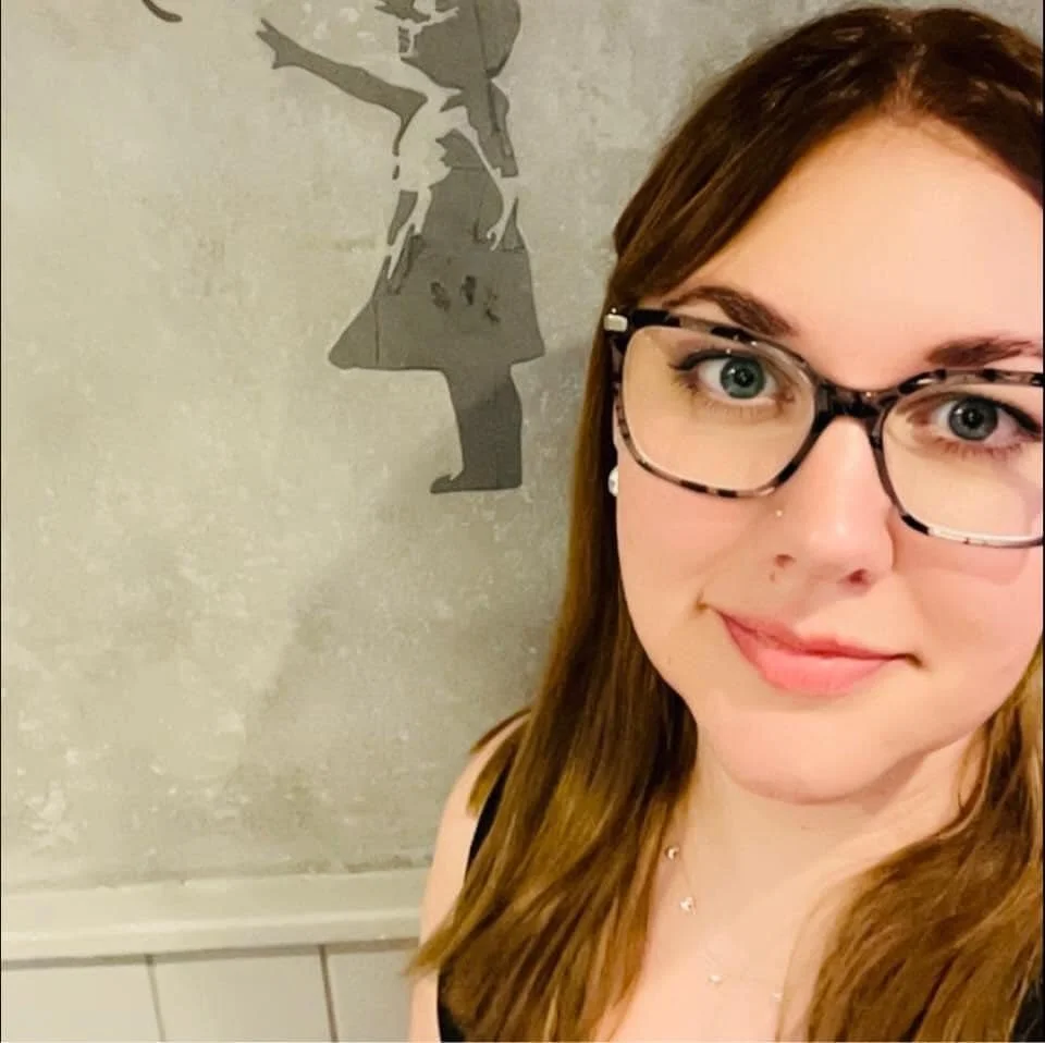 A young woman with long brown hair and glasses in front of a bansky.
