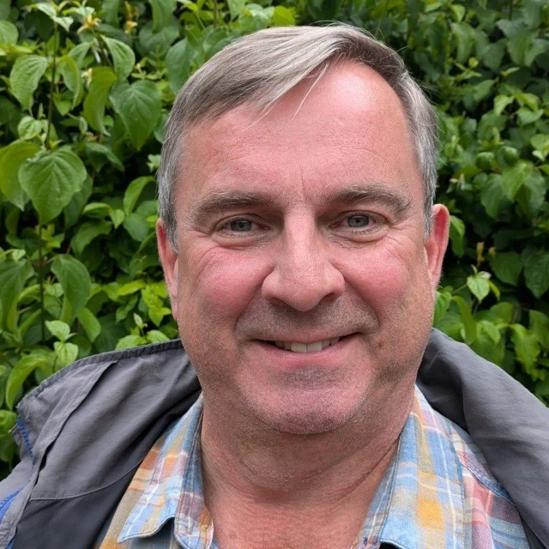 A man smiling outdoors with green bushes in the background.