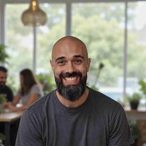 A smiling man with a bald head and beard sitting indoors in front of large windows with a green cafe background.