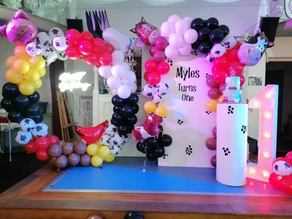 Balloon arch with cow print, yellow, red, black, and white balloons, around a birthday sign that reads "Myles Turns One." There is a large illuminated number one, a tiered birthday cake, and party decorations.