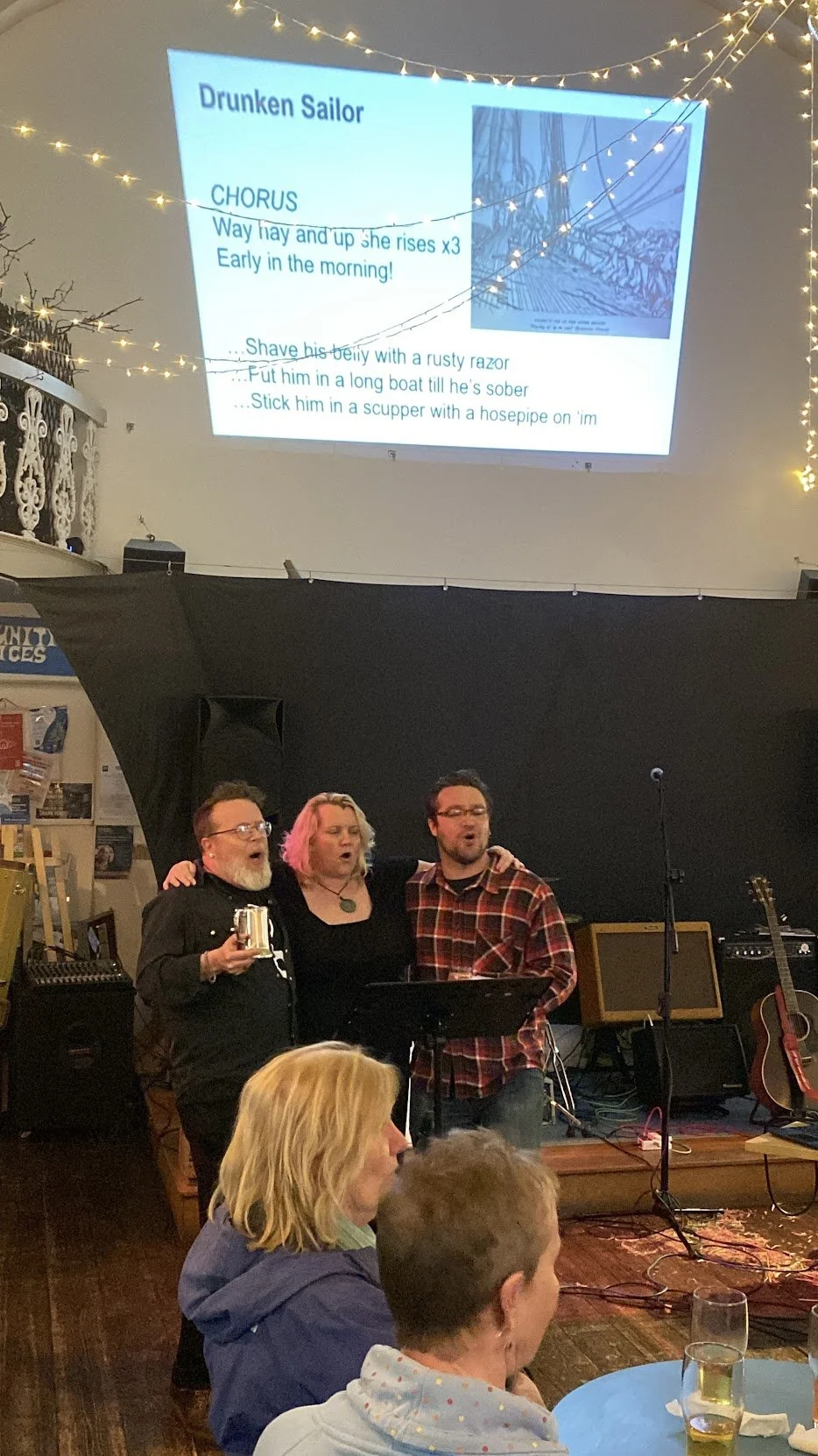 Three people singing together arms round each other on stage during a karaoke night, the large screen behind them displaying song lyrics and an image, in our beautiful café space.