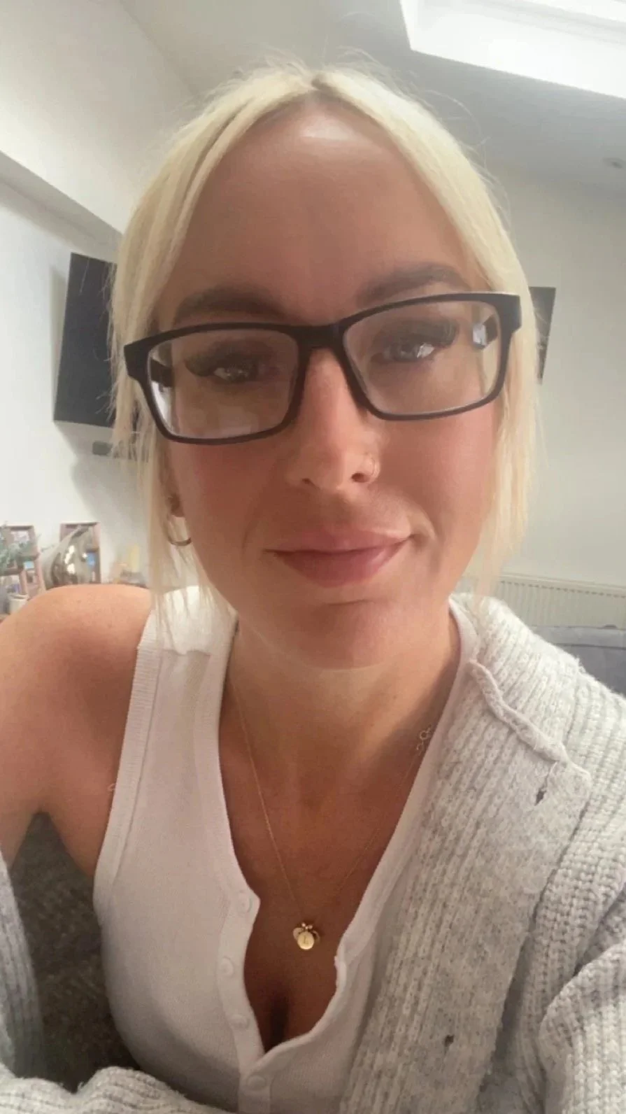 A woman with blonde hair, glasses, and a nose ring smiling in a close-up selfie. She is wearing a white top, a grey cardigan, and a gold necklace with a small round pendant.