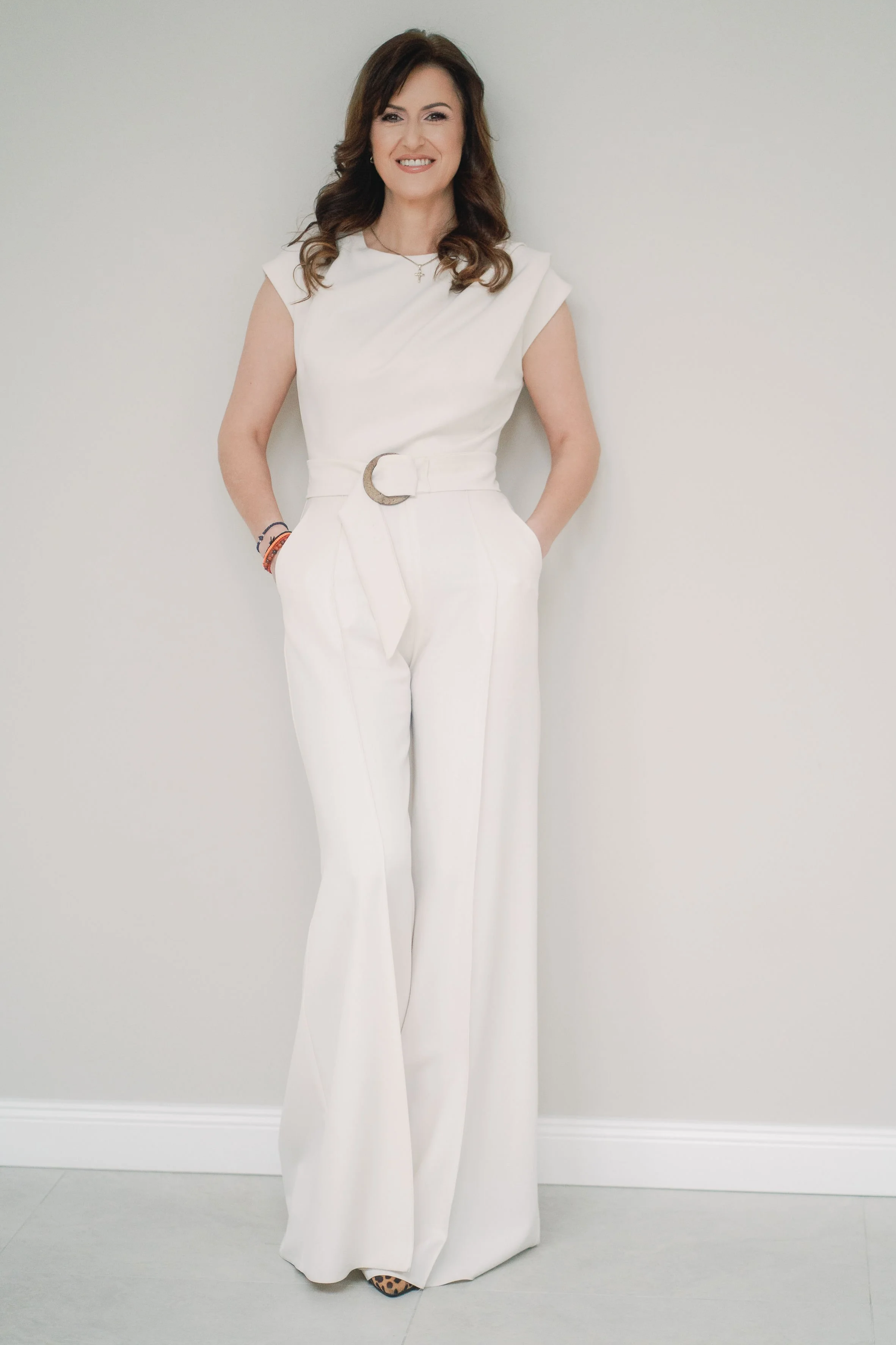 A woman dressed in a white sleeveless jumpsuit with a belt, standing against a plain white wall background, smiling with her hands in her pockets.