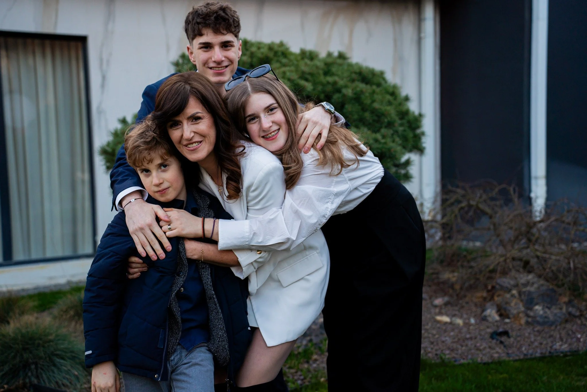 A family of five enjoying a group hug outdoors in front of a house with large windows and bushes.