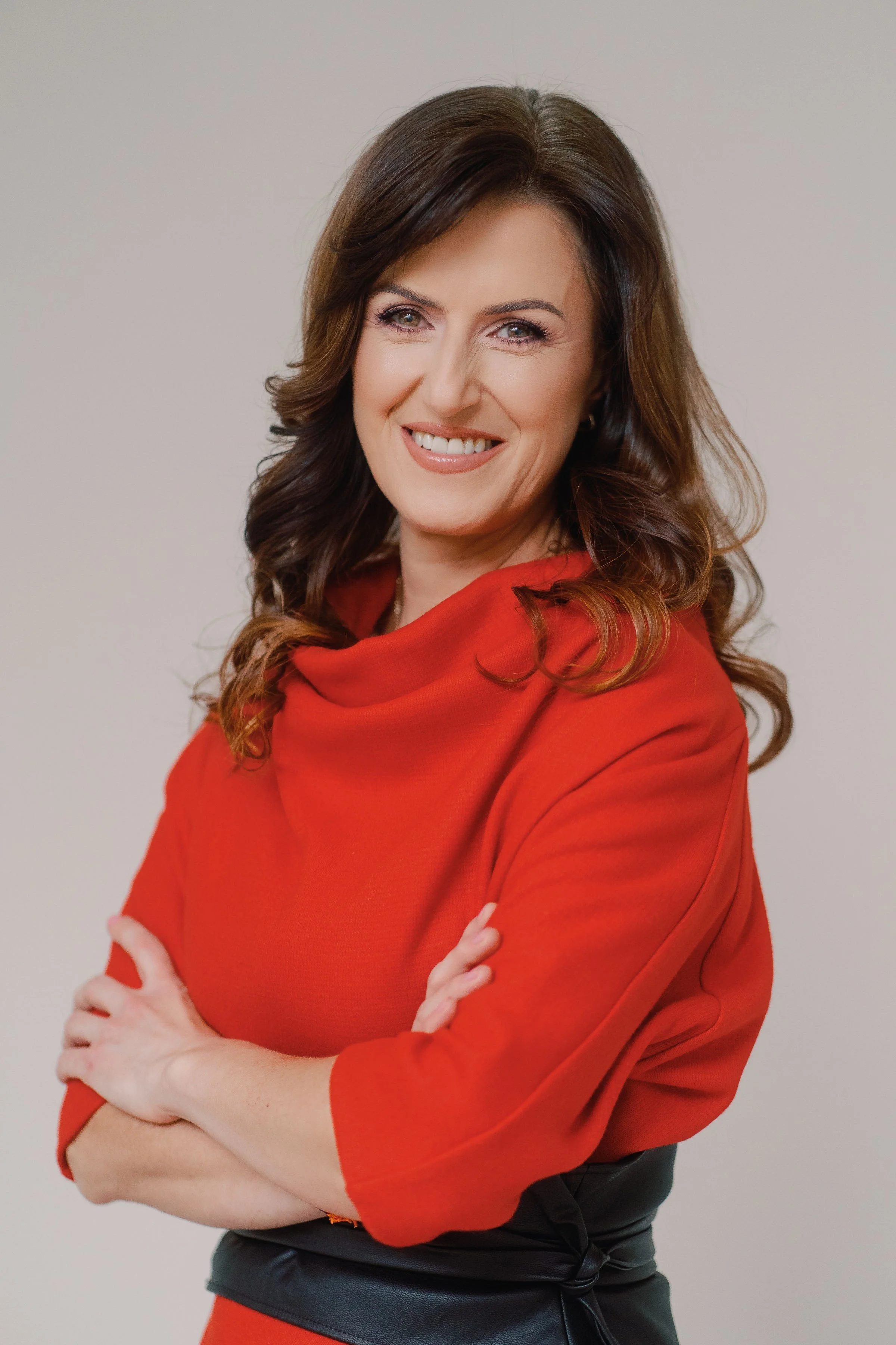 A woman with wavy brown hair and a bright smile, wearing a red top and black pants, standing with her arms crossed against a plain white background.