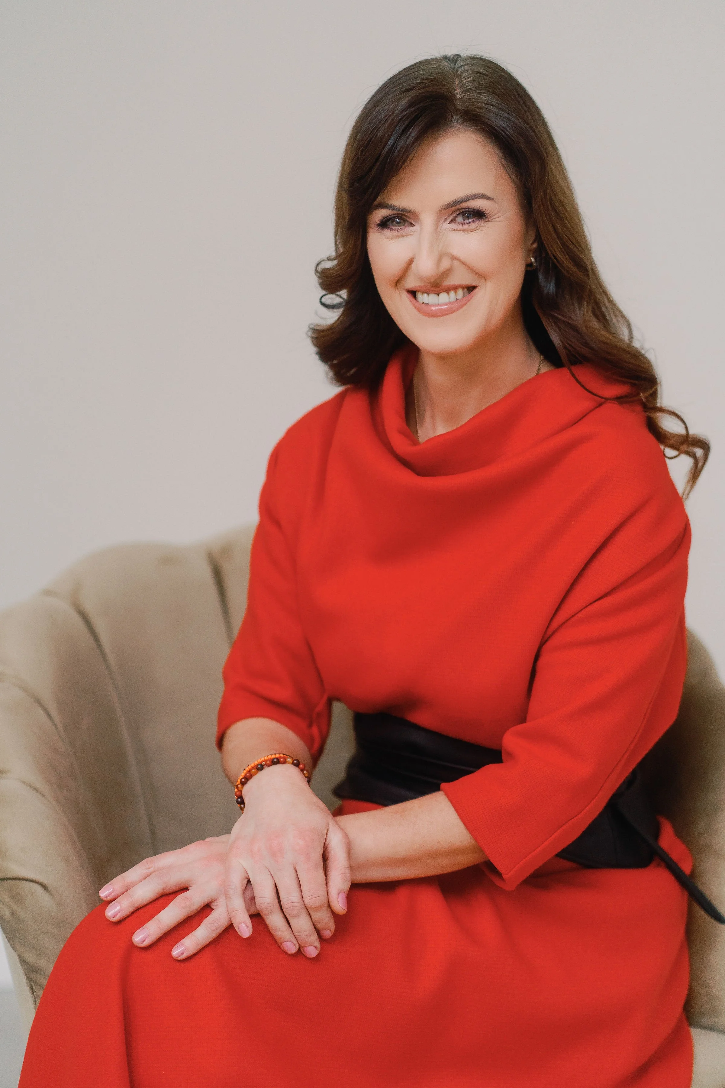 A woman with brown hair wearing a red dress, sitting on a beige sofa, smiling, with her hands resting on her lap, wearing bracelets.