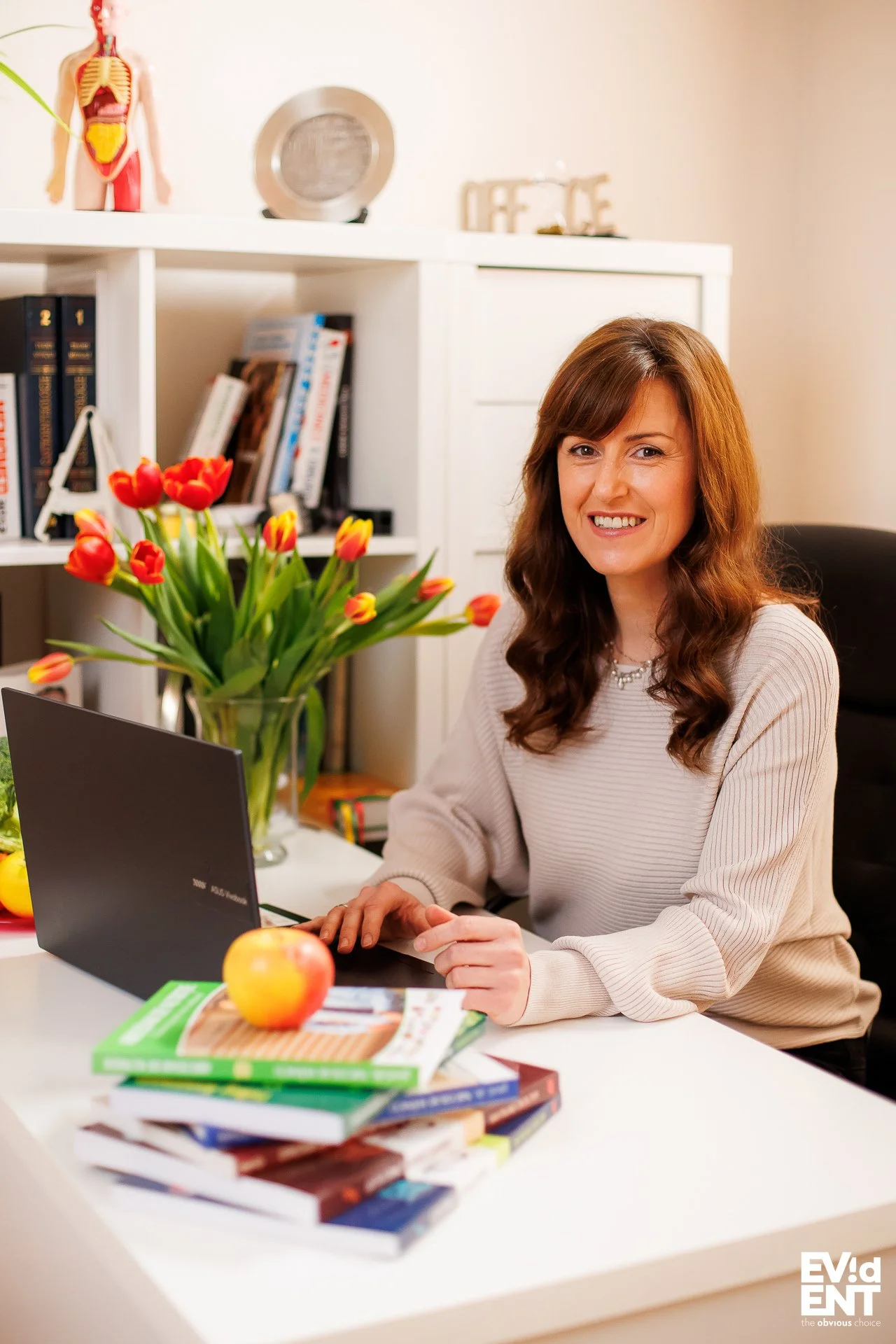 A woman sitting at a desk with a laptop, a vase of red and yellow tulips, a stack of books, and an apple, in a room with bookshelves and decorative items.