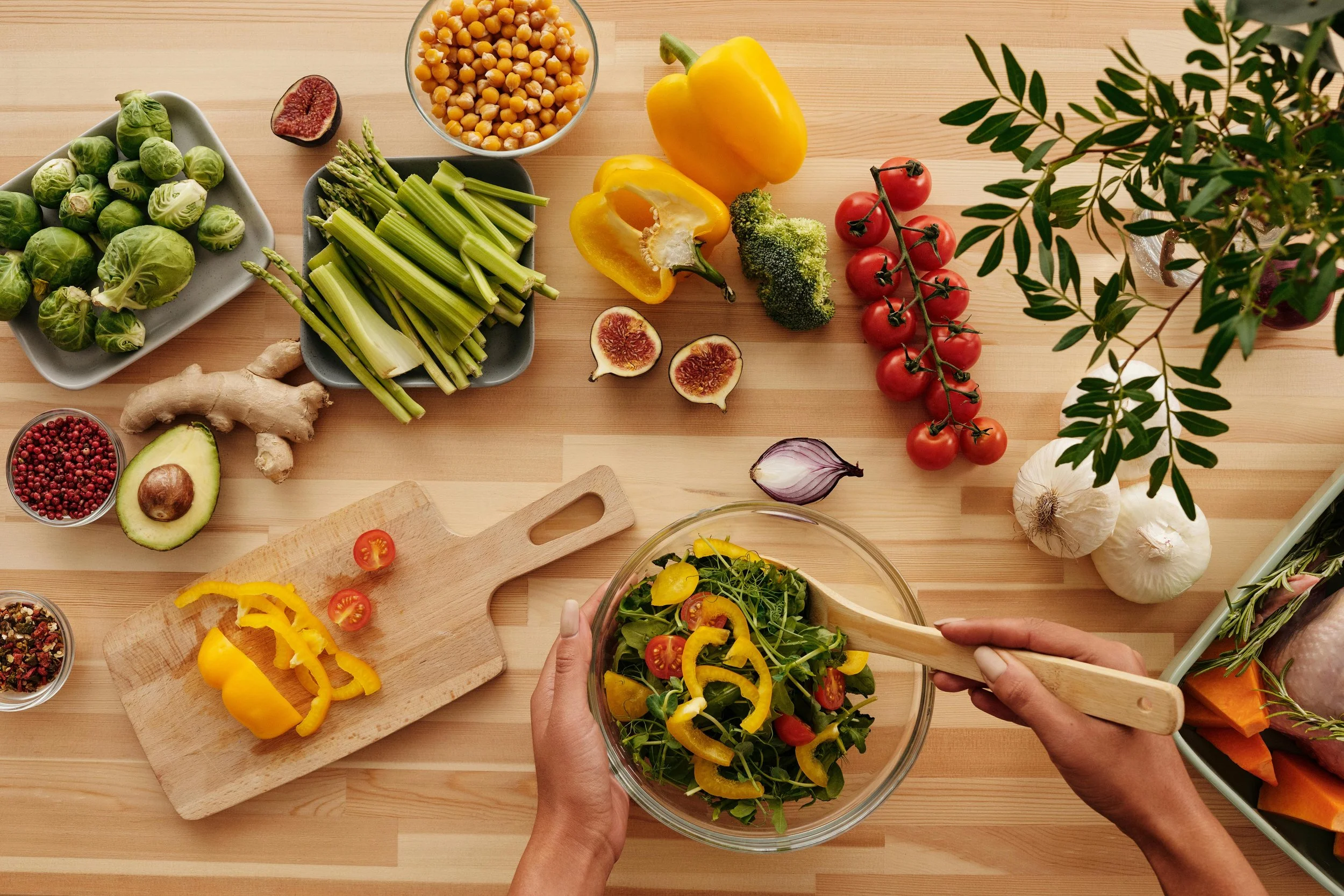 A person preparing a fresh vegetable salad with various vegetables including yellow bell peppers, tomatoes, broccoli, celery, Brussels sprouts, garlic, figs, ginger, and a bowl of chickpeas on a wooden table. Someone is tossing sliced yellow bell peppers and cherry tomatoes into a glass bowl of salad.