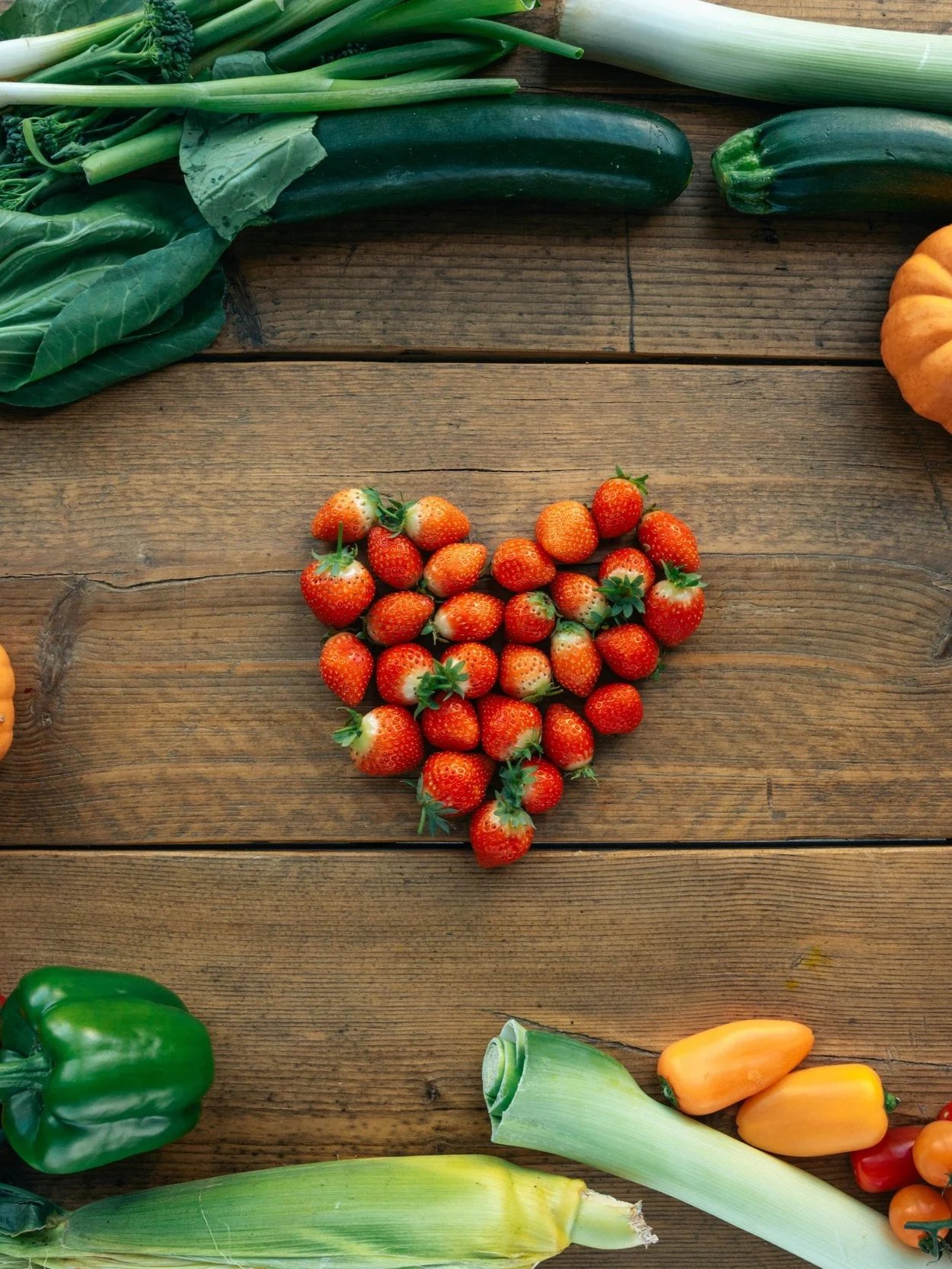 Various fresh vegetables including strawberries arranged in a heart shape, with zucchini, green bell pepper, yellow cherry peppers, and green onions on a rustic wooden surface.