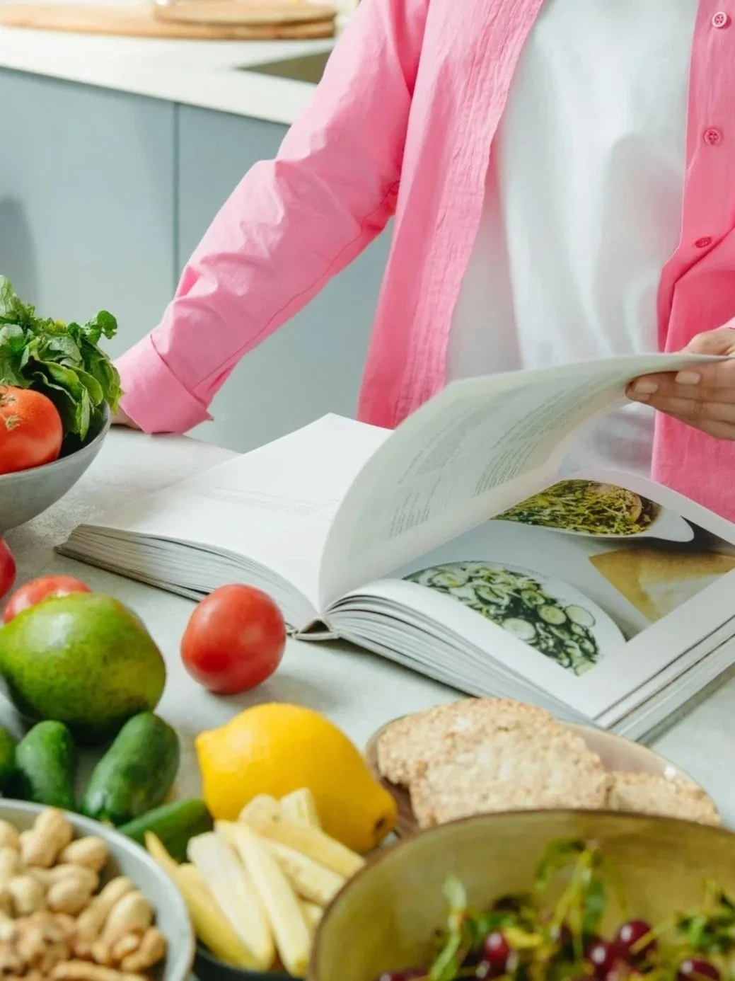 A person in a pink shirt looking at a cookbook in a kitchen with fresh vegetables, fruits, and nuts on the counter.