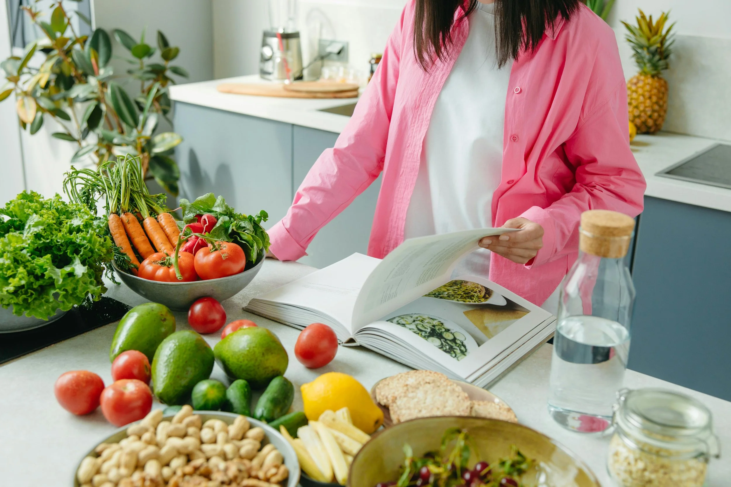 A woman in a pink shirt preparing a variety of fresh vegetables and fruits on a kitchen countertop, including carrots, tomatoes, green apples, lemons, and leafy greens, with an open book in front of her.