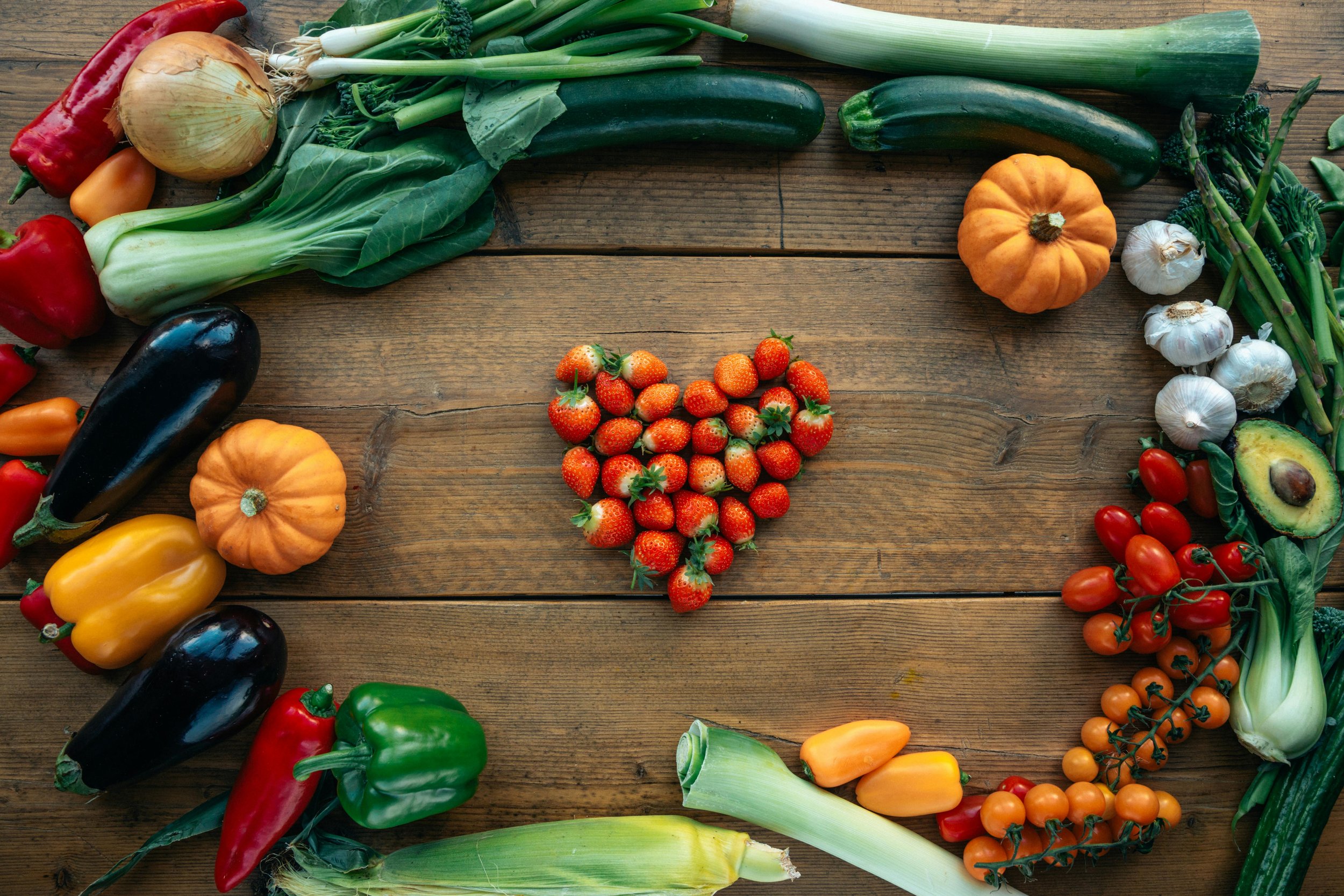 Assorted fresh vegetables arranged in a circle on a wooden surface, including strawberries in the center, and zucchini, eggplant, peppers, garlic, onion, green onions, cherry tomatoes, leek, broccoli, avocado, and winter squash.