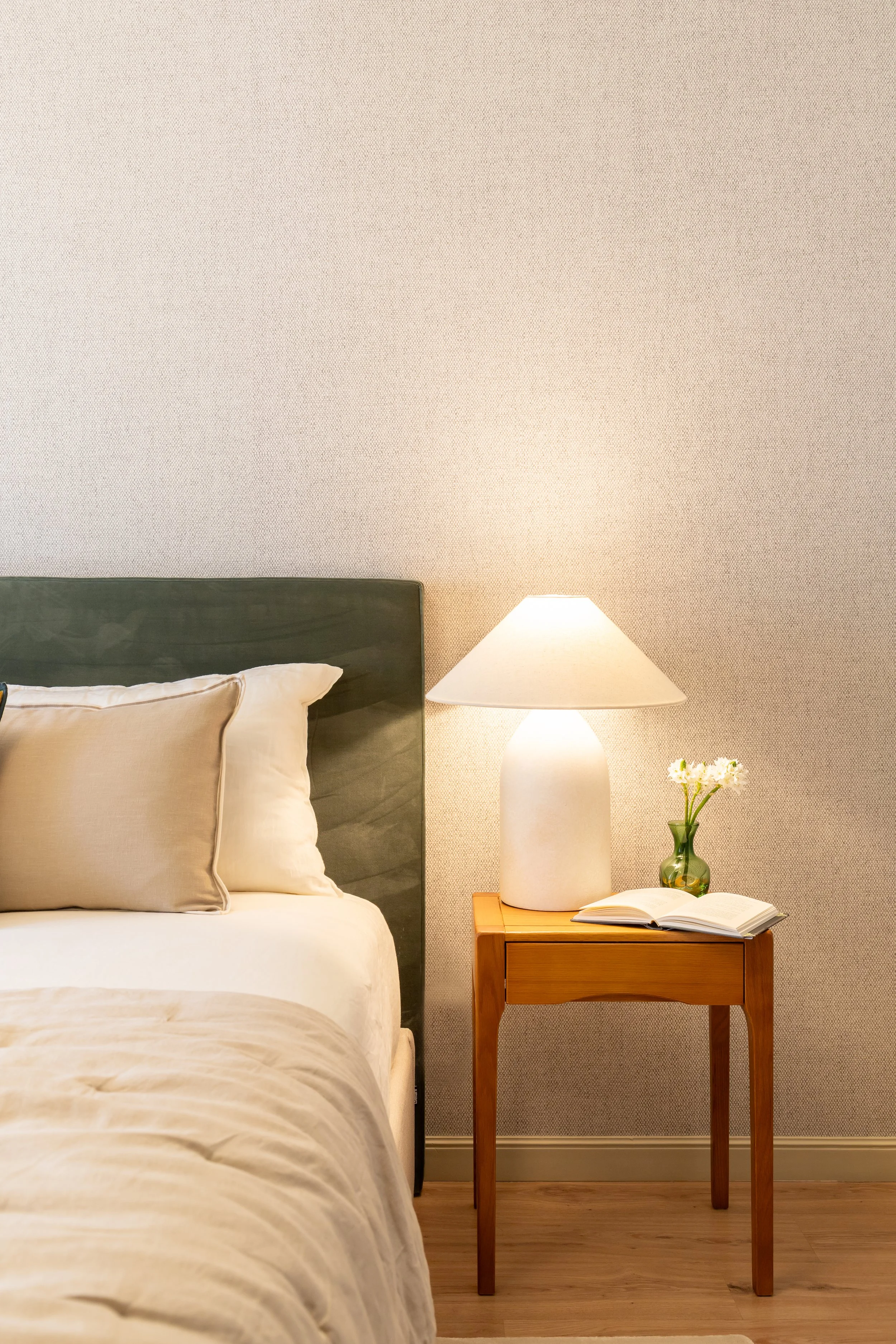 Quarto com roupa de cama branca, mesa de apoio em madeira e flores