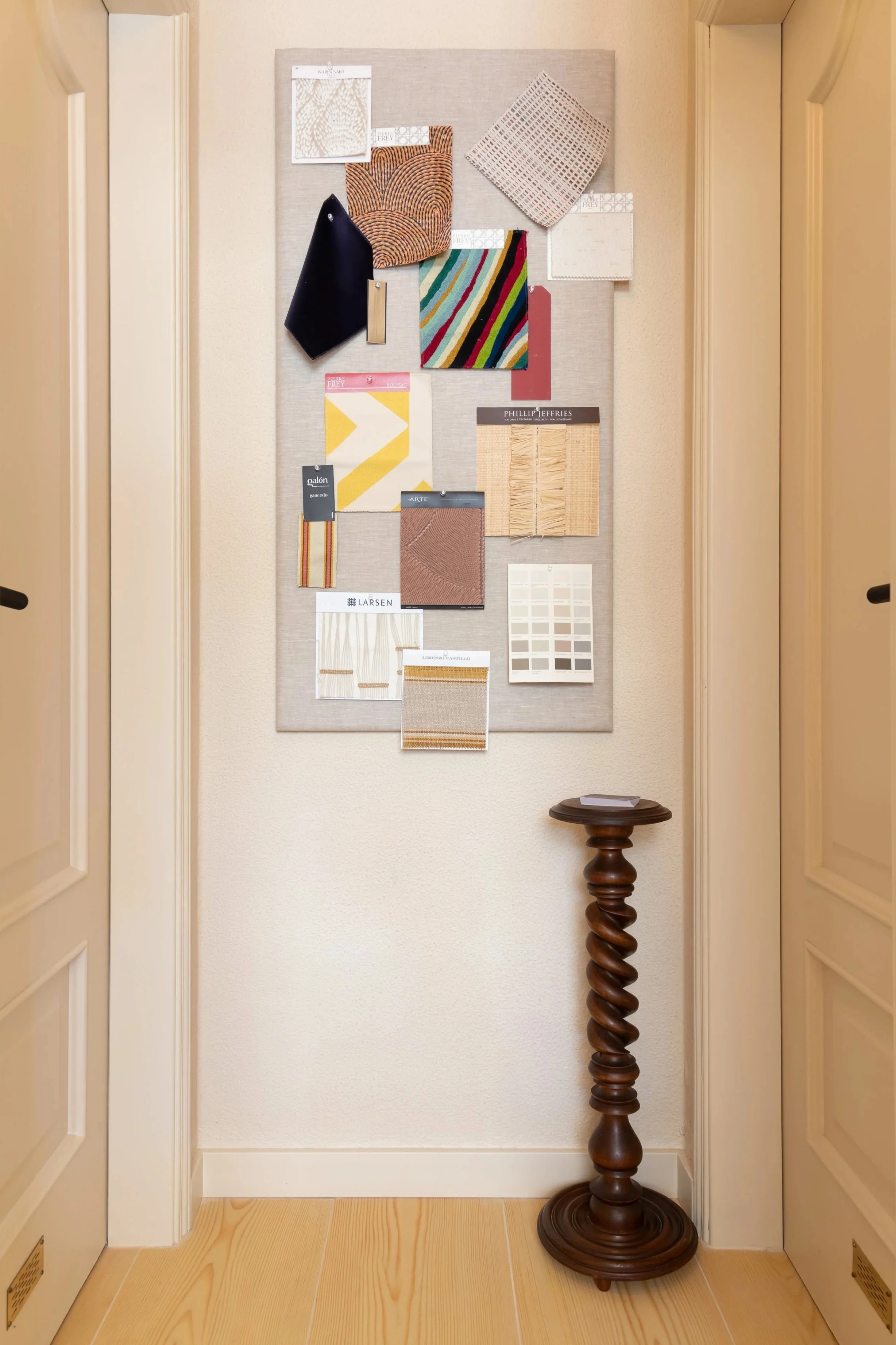 Corrediça de parede com amostras de tecidos e materiais de design, decorada com uma moldura de tecido, amostras de papel, um pequeno quadro, e outros acessórios, além de um lustre de madeira no chão ao lado direito.