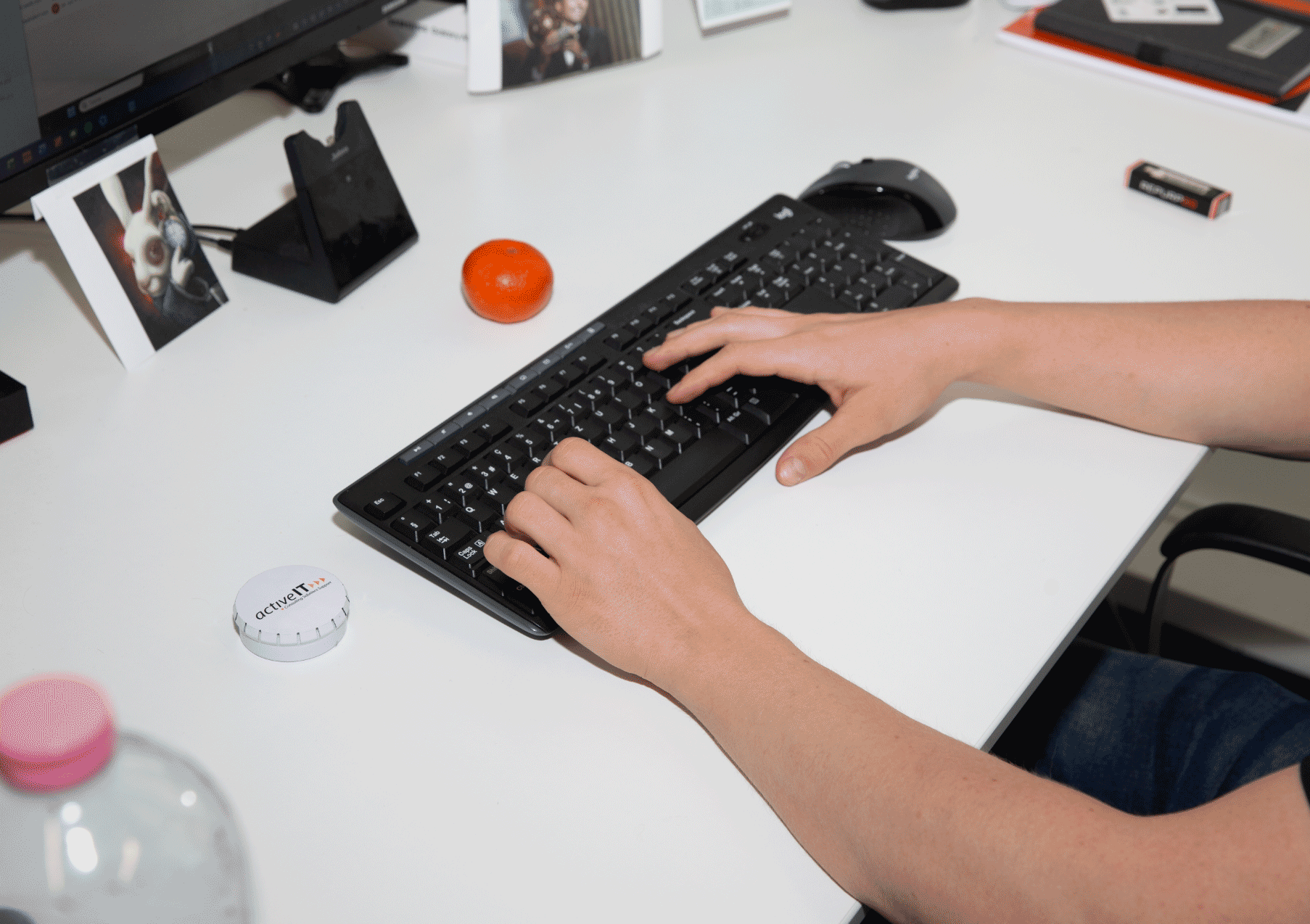 Person tippt auf einer schwarzen Computertastatur auf einem weißen Schreibtisch, auf dem verschiedene Gegenstände wie eine orange Kugel, eine weiße Dose mit dem Aufdruck 'activeIT', eine Computermaus, eine Kamera, ein Mikrofon, ein Foto und ein Akku liegen.