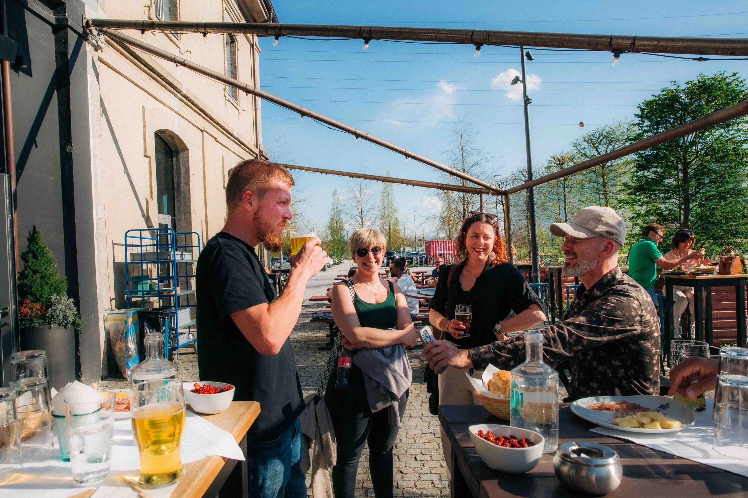 Menschen, die bei einem Treffen im Freien essen und trinken, während sie an einem Tisch mit Essen und Getränken sitzen und sich unterhalten, bei schönem Wetter in einer Stadtumgebung.