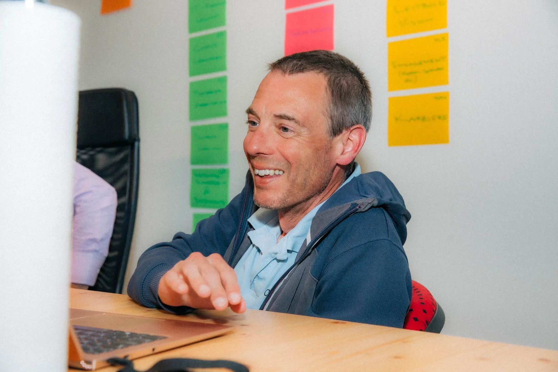 Ein lächelnder Mann sitzt an einem Tisch, neben einem Laptop, in einem Büro mit bunten Sticky Notes an der Wand.