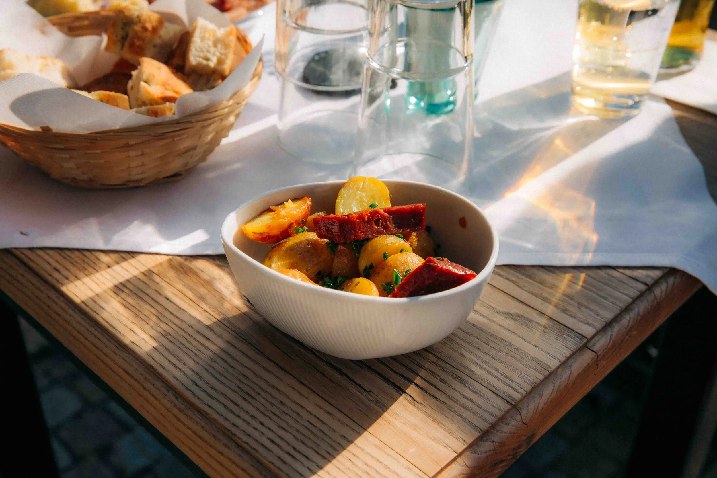 Blick auf eine Schüssel mit Kartoffelsalat und Grillwürstchen, daneben ein Korb mit Brot und leere Gläser auf einem Holzbrett, Schatten und Sonnenlicht auf dem Tisch.