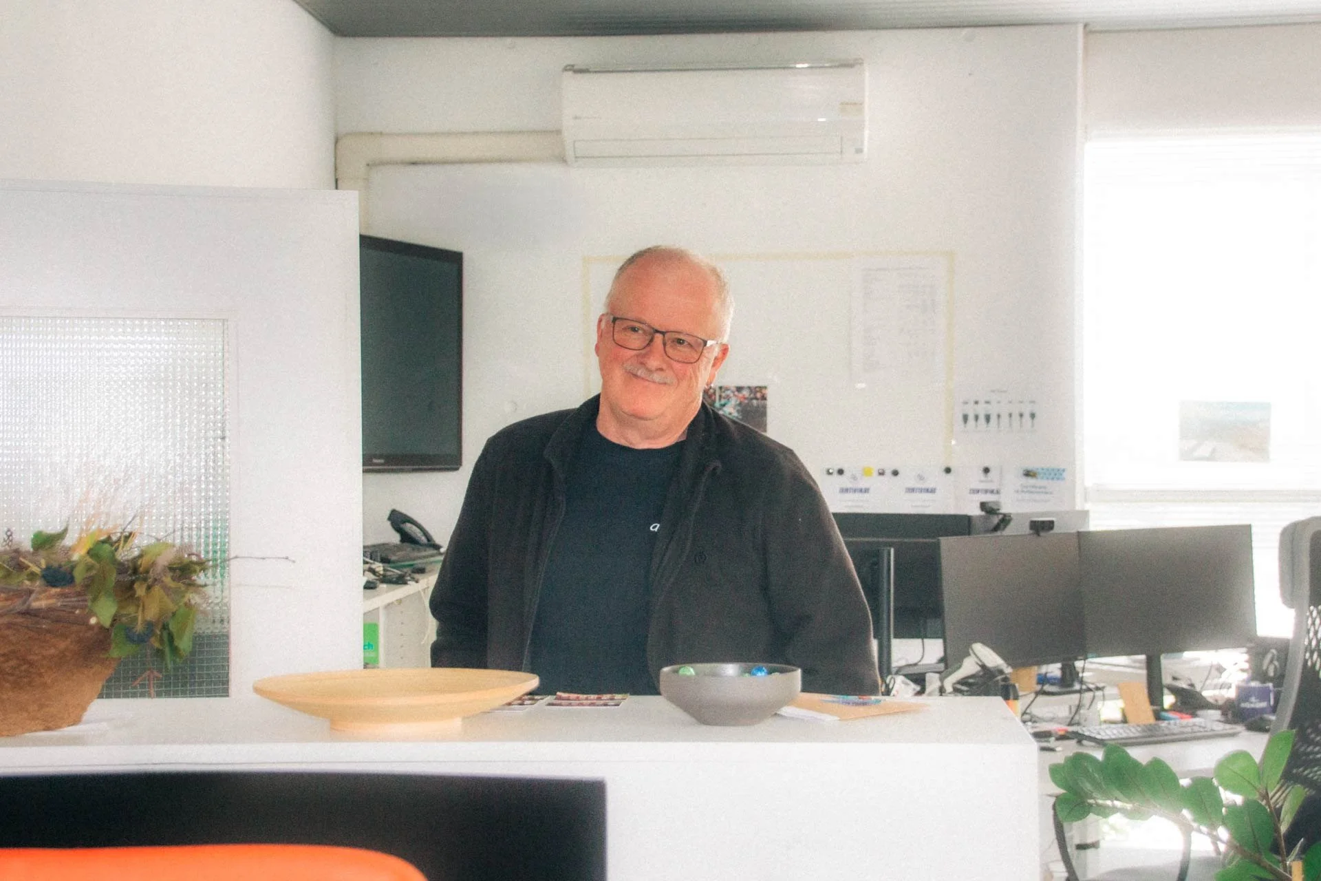 Ein älterer Mann mit Brille und dunklem Hemd steht im Büro, lächelt und schaut in die Kamera, im Hintergrund sind nummerierte Whiteboards, Bildschirme und Arbeitsplätze zu sehen.