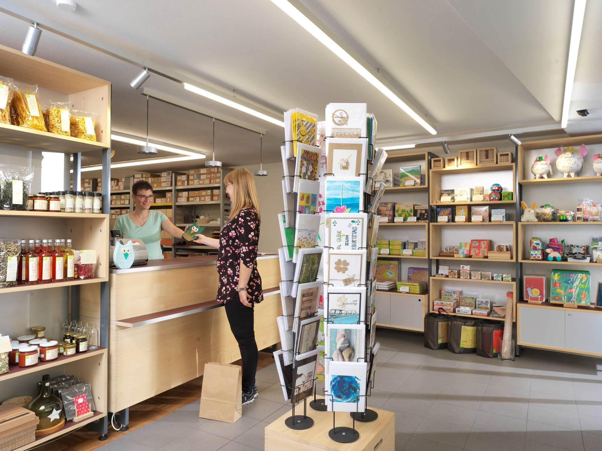 Ein Laden mit Holzregalen, in denen Produkte wie Lebensmittel, Getränke, Bastelmaterialien und Dekorationen stehen. Zwei Frauen am Ladentisch im Gespräch, eine von ihnen überreicht der anderen ein Produkt. Im Hintergrund sind weitere Regale mit bunten Artikeln und Grußkarten zu sehen.