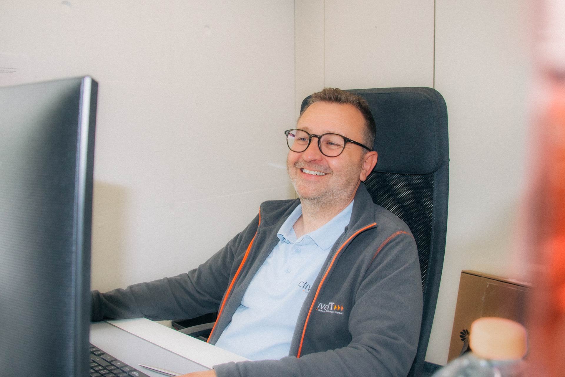 Ein Mann mit Brille sitzt am Schreibtisch vor einem Computerbildschirm in einem Büro und lächelt.