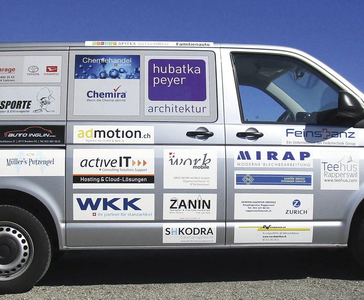 Seitenansicht eines weißen Fahrzeugs mit mehreren Firmenwerbungsschildern auf den Türen, darunter Chemiehandel, Architektur, Hosting, Möbel, Sanitäre Dienste, Versicherungen und Handwerksbetriebe, vor einem blauen Himmel.